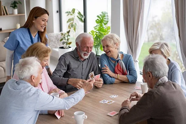 Group of elderly people playing cards at a table with a caregiver supervising in a bright room.