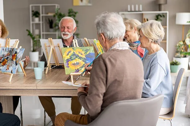 Group of elderly people in a painting class, sitting around a table with easels and colorful paintings, smiling and enjoying art activity in a bright room.