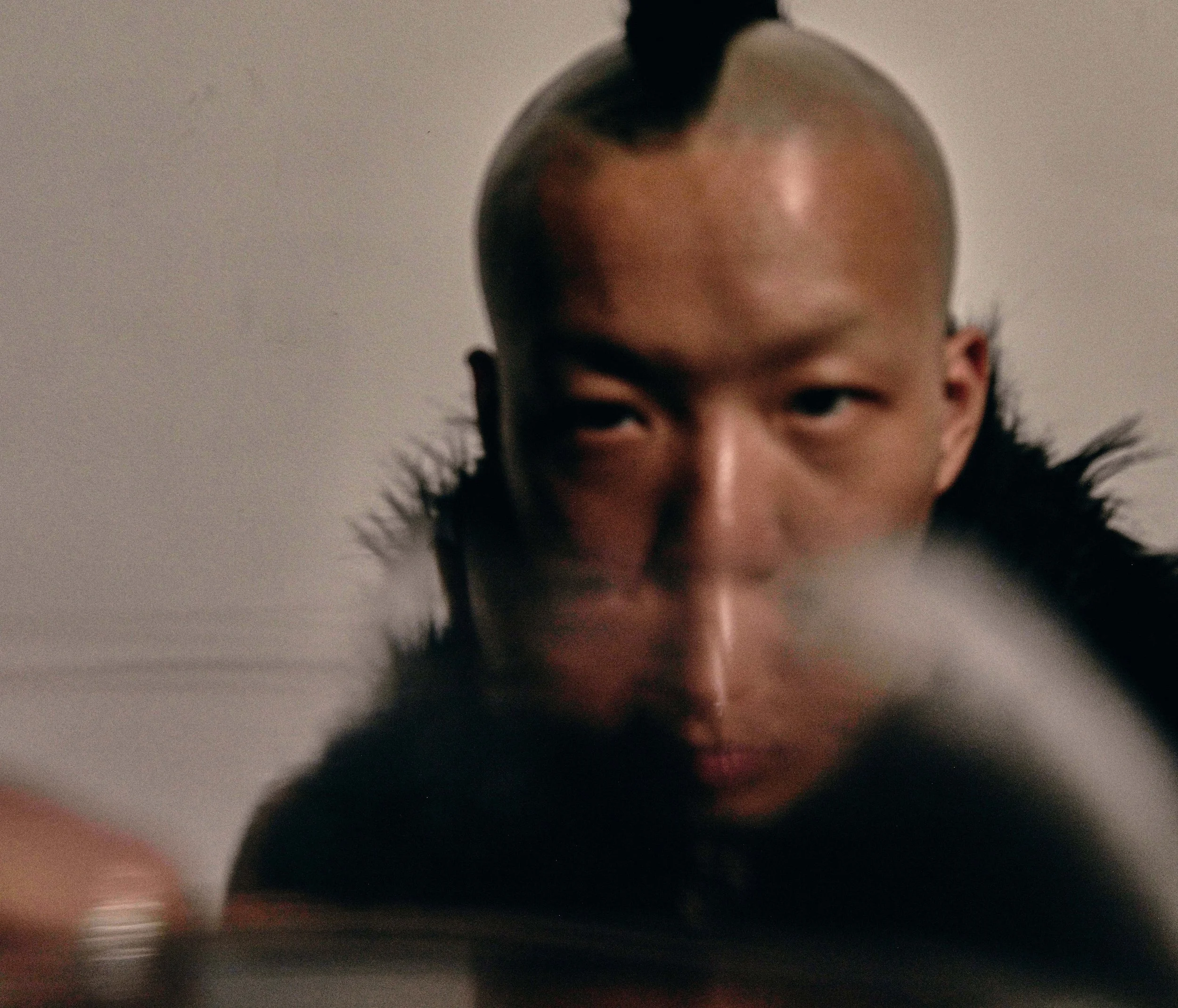 Close-up of a person's face with an unusual bald head, partially visible, with dark makeup and black hair styled in a high bun, against a plain background.