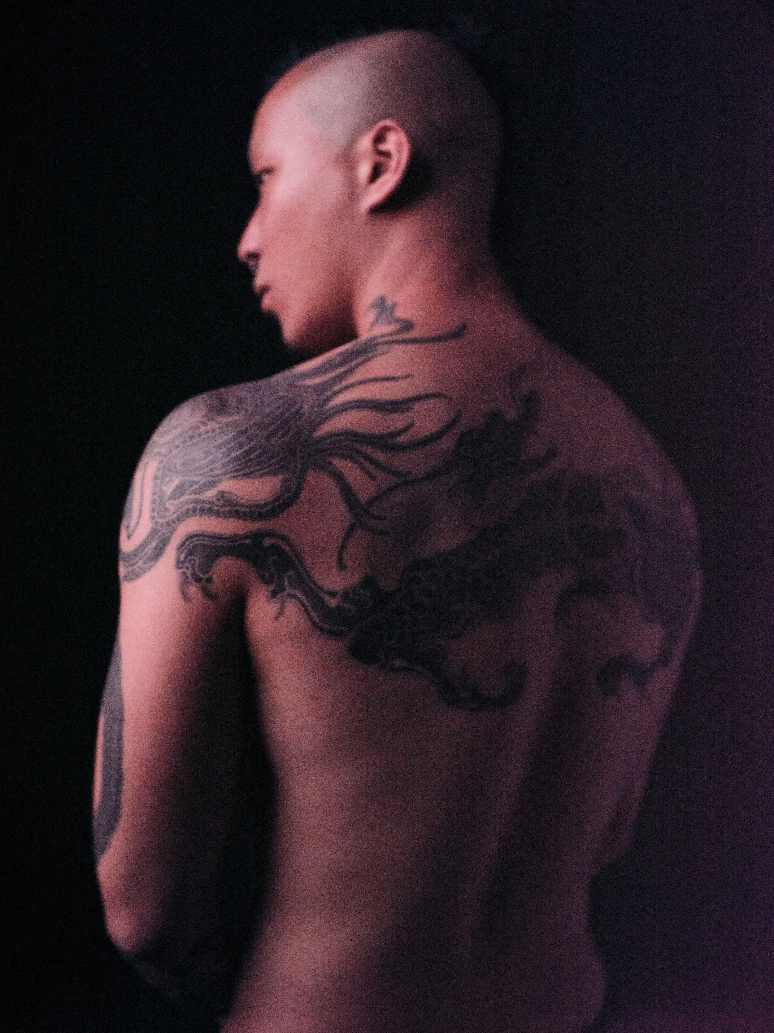 Side view of a person with a buzz cut hairstyle, showing their neck and shoulder, with tattoos on the upper arm and back, against a dark background.