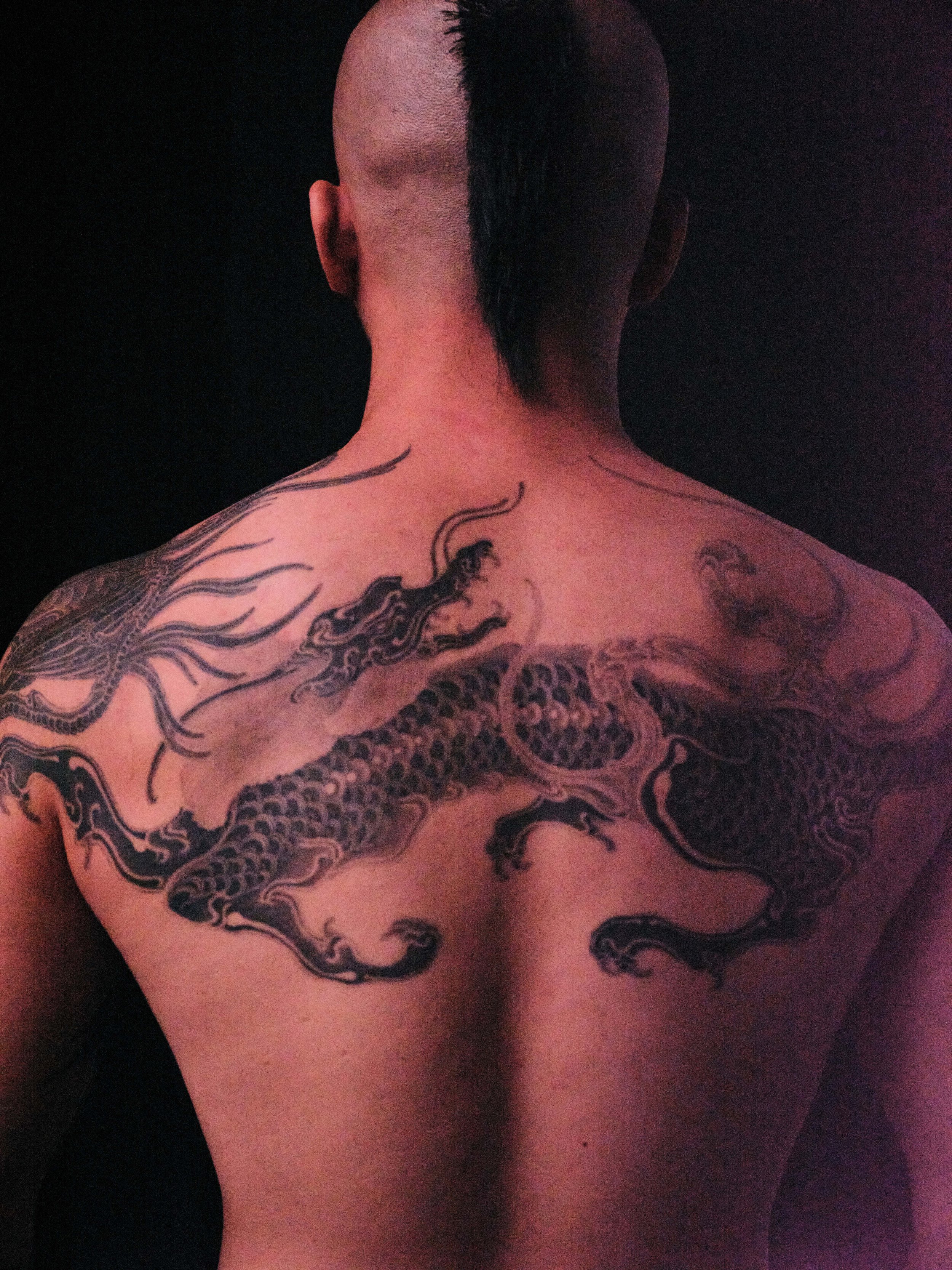Back view of a person with a mohawk hairstyle, featuring shaved sides and a longer strip of hair in the middle. The person has tattoos on shoulders and upper back, including a large dragon design. Shot by Photographer Ian O'Hara