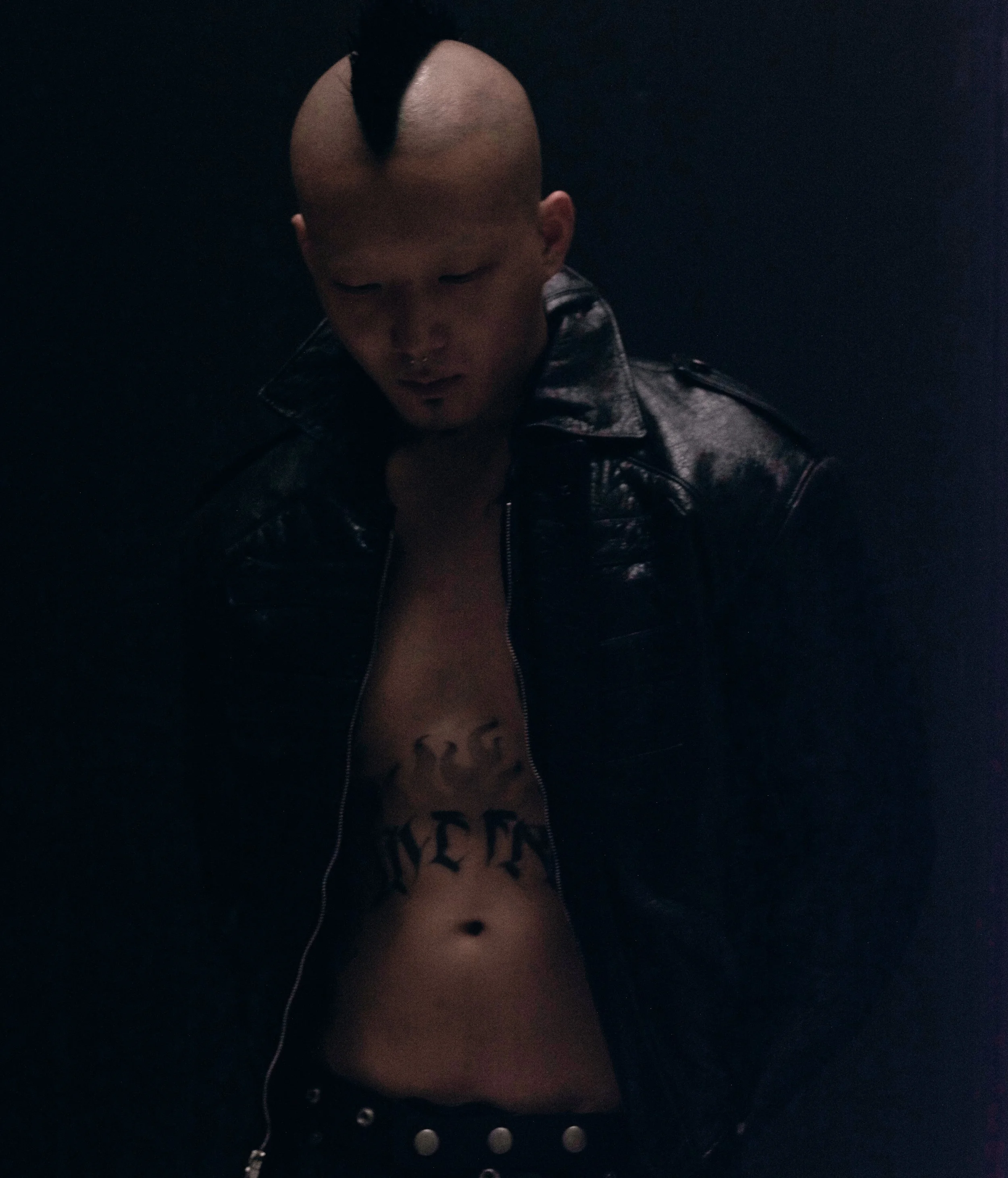 A person with a shaved head and a black Mohawk hairstyle, wearing a black leather jacket, and black top with text, against a dark background.