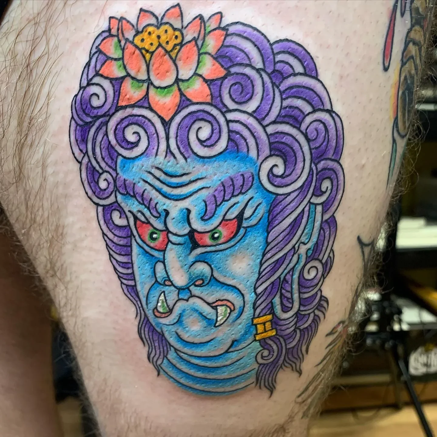 Colorful tattoo of a fierce blue and purple demon face with red eyes and sharp fangs, adorned with an orange lotus flower on the forehead and swirling purple hair.