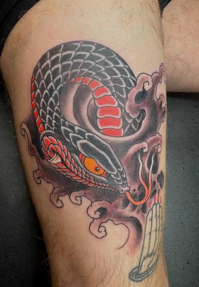 Tattoo of a coiled snake with black and gray scales, orange accents, and yellow eyes, surrounded by stylized pink and black clouds.