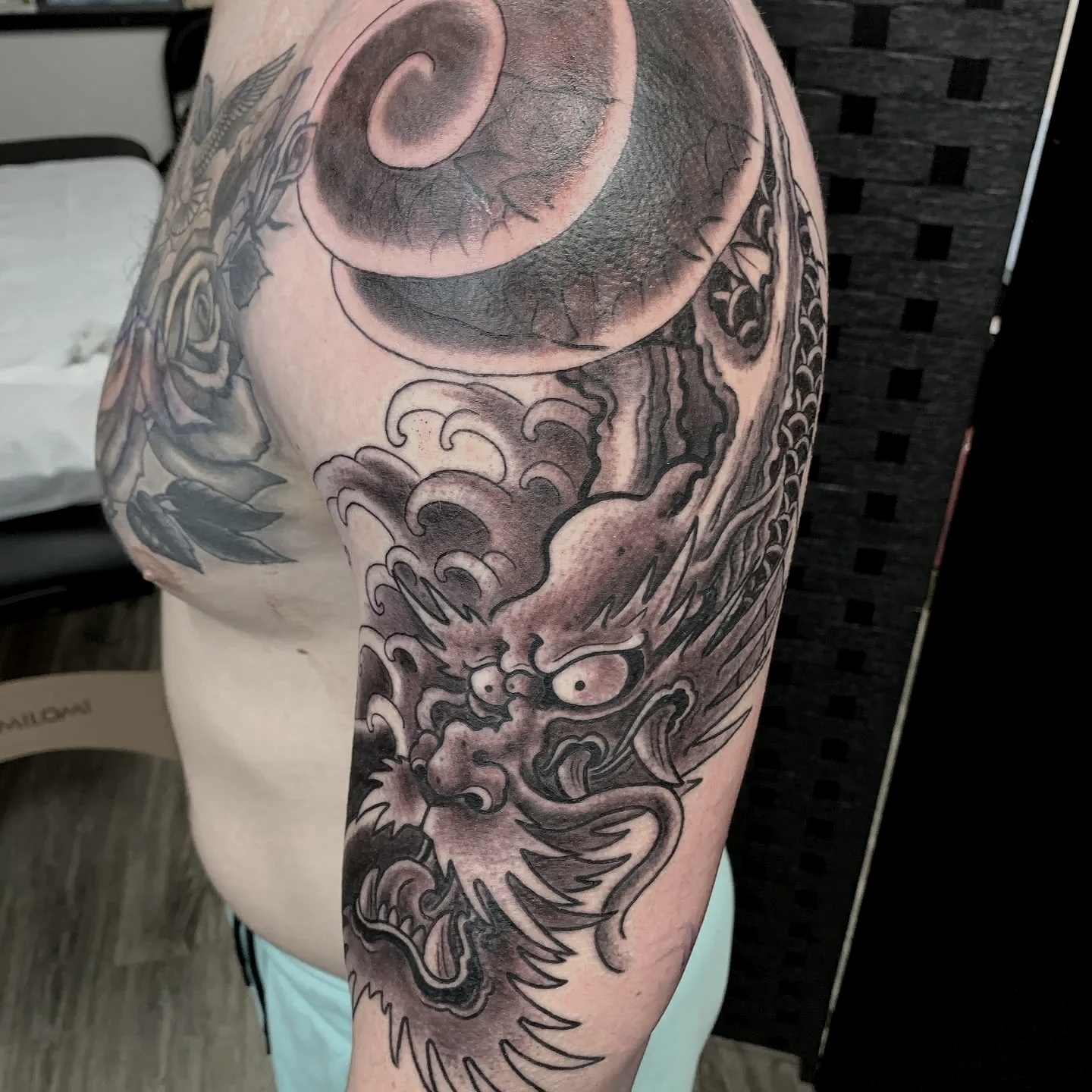 Dragon half sleeve — black and grey with traditional Japanese wave and swirl background, large shoulder cap flowing into forearm. Bold outline, heavy whip shade, built for long-term hold.
