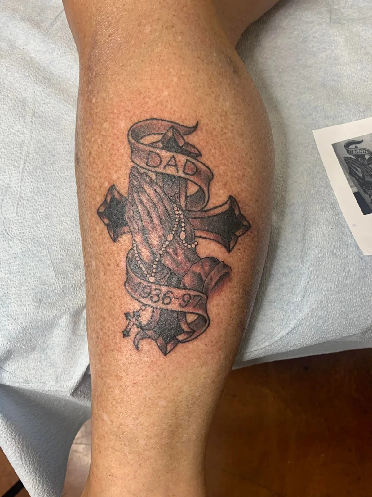 Custom memorial tattoo featuring praying hands with rosary over a bold cross, wrapped with banners reading DAD and 1936-97. Black and gray tribute by Brett Semrick