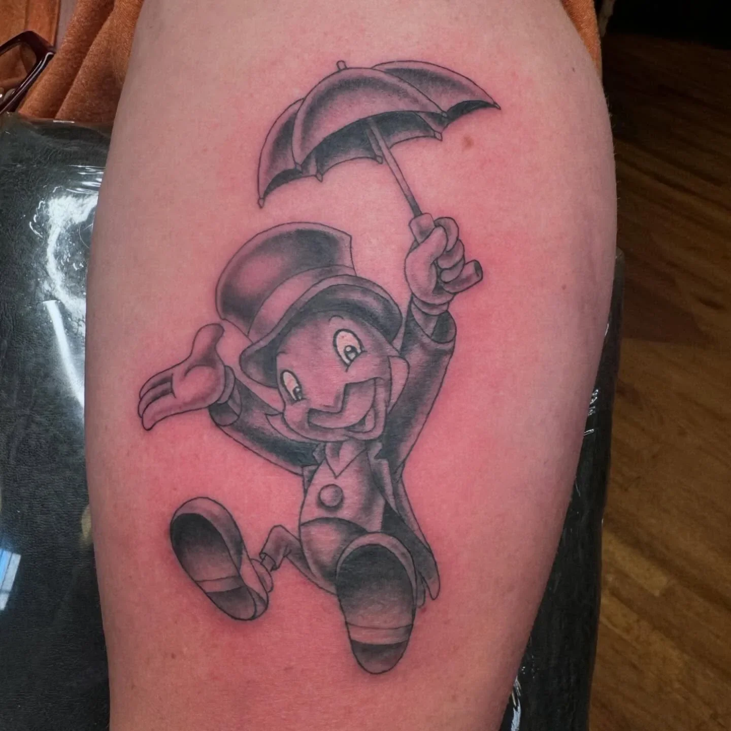 Charming black and gray Jiminy Cricket tattoo from Pinocchio wearing top hat and holding umbrella mid-jump. Soft shaded cartoon realism by Brett Semrick in San Diego.