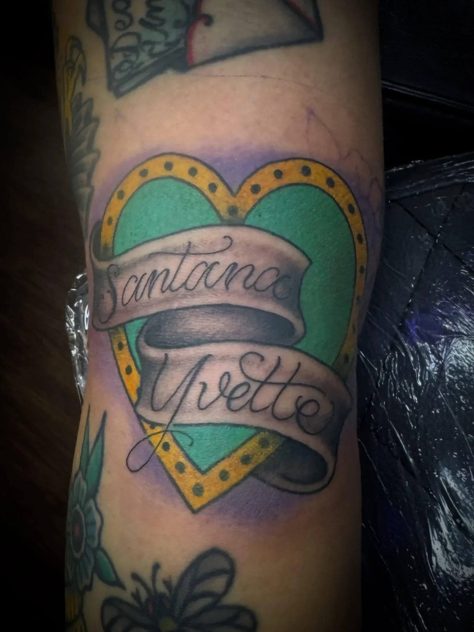 : Color traditional heart tattoo with Santana Yvette script banners - green heart with gold dotted border and purple shading. Classic love name tattoo by Brett Semrick.
