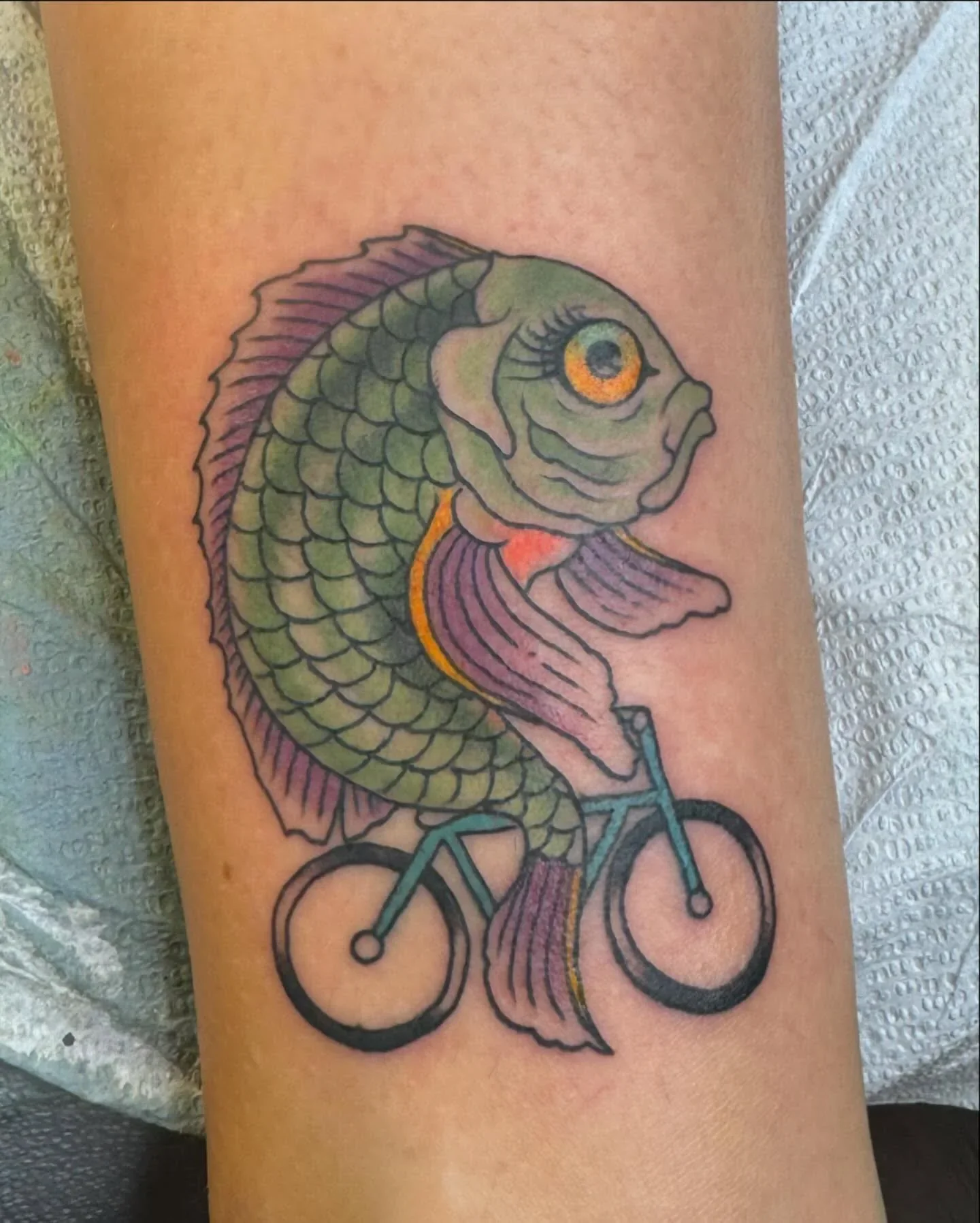 Whimsical traditional tattoo of a green scaled fish riding a teal bicycle. Funny and surreal color traditional tattoo by Brett Semrick.