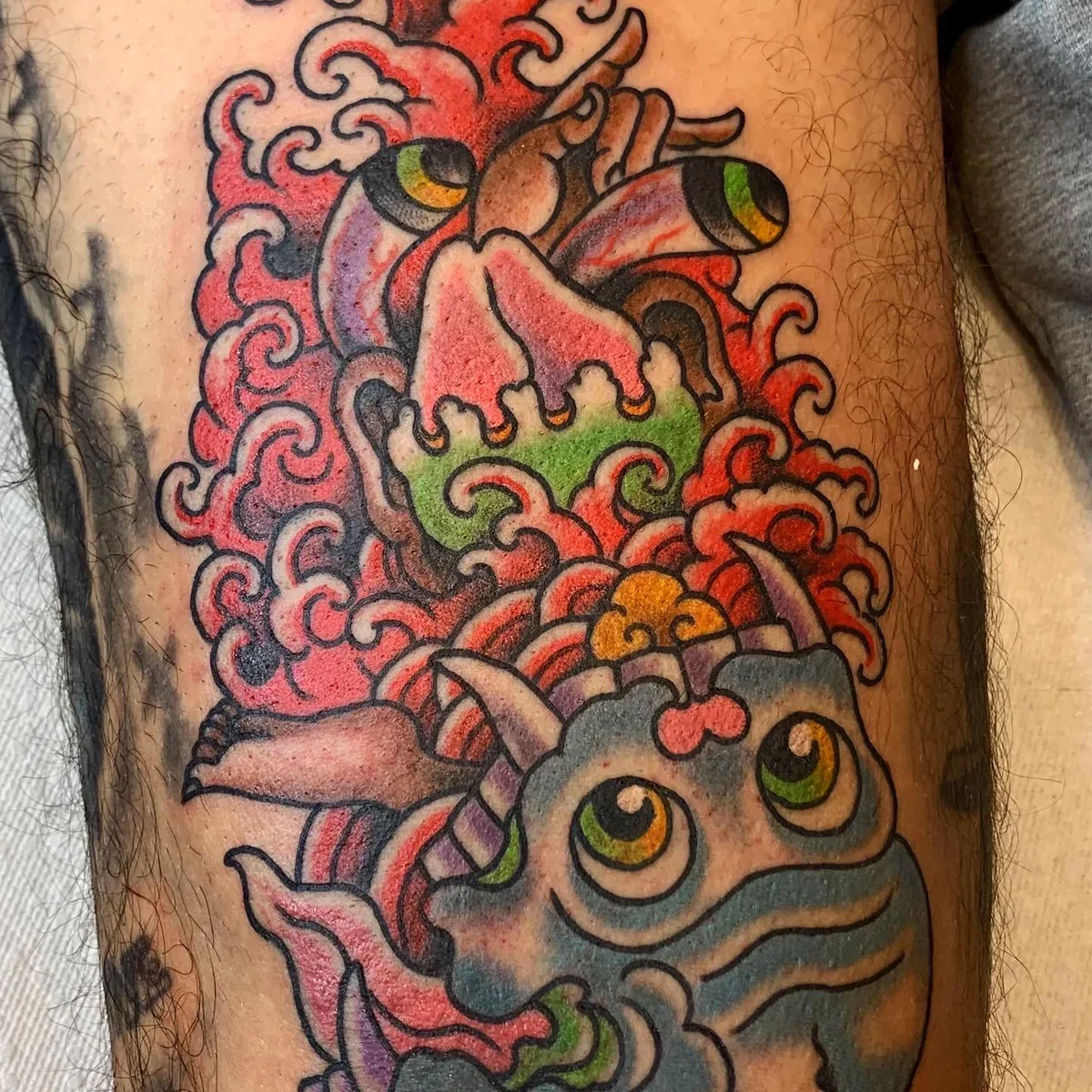 Colorful tattoo of a whimsical dragon with green eyes, surrounded by red, pink, orange, yellow, purple, and black swirling clouds and flames.