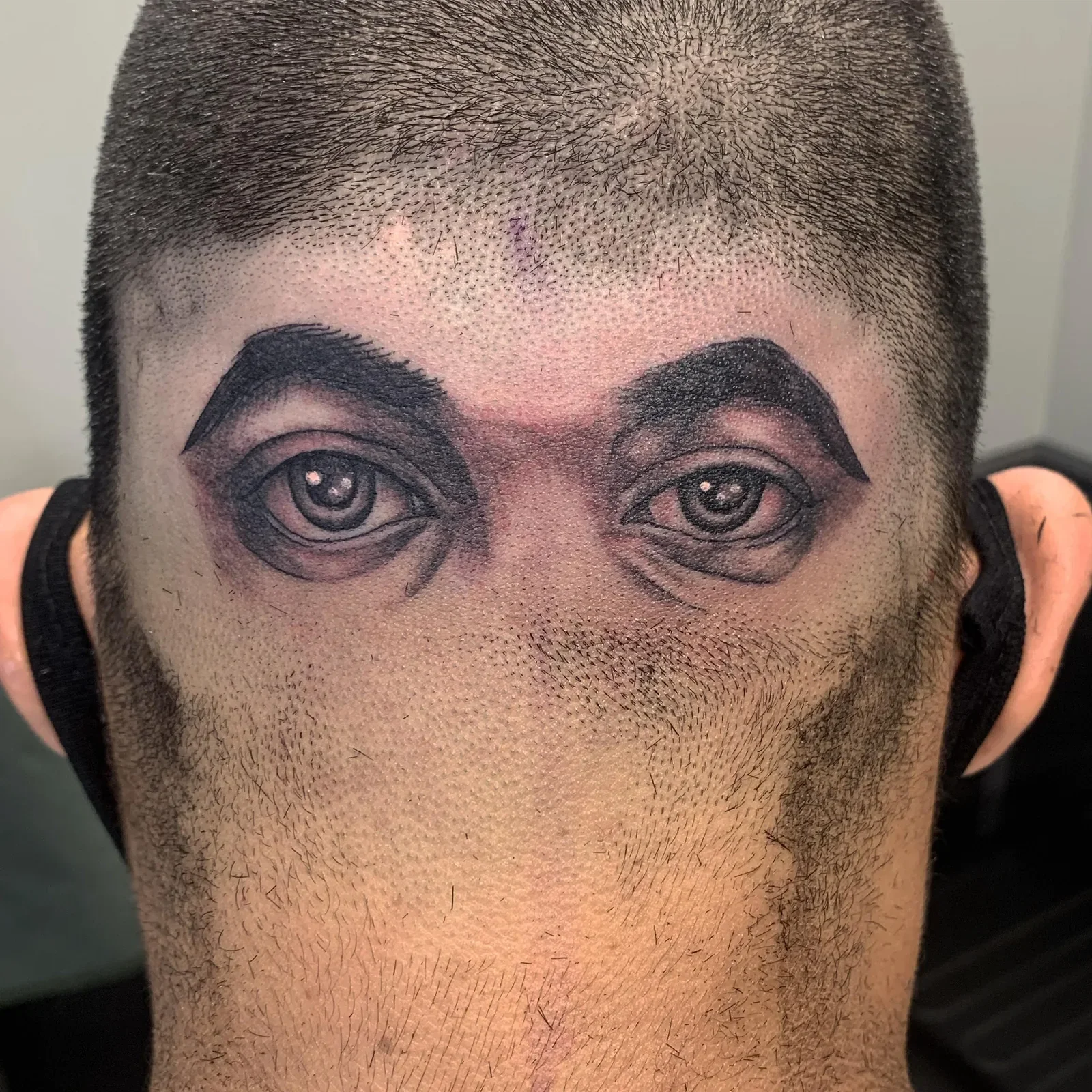 Trompe l'oeil realistic eyes on back of shaved head — black and grey realism with full eyebrows and iris detail.
