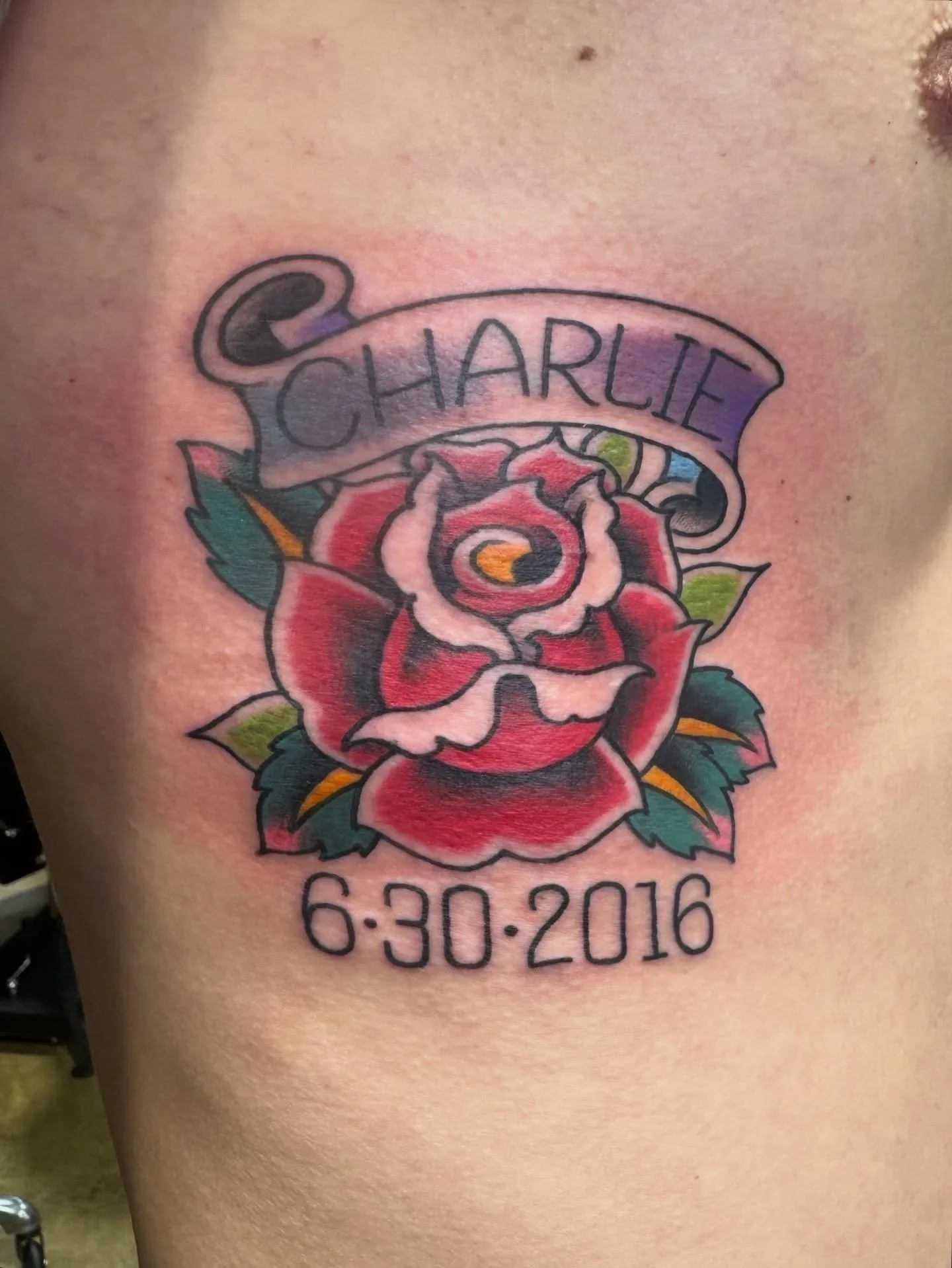 Color traditional red rose tattoo with Charlie banner and date 6-30-2016. Classic memorial style with green leaves and purple banner shading by Brett Semrick.
