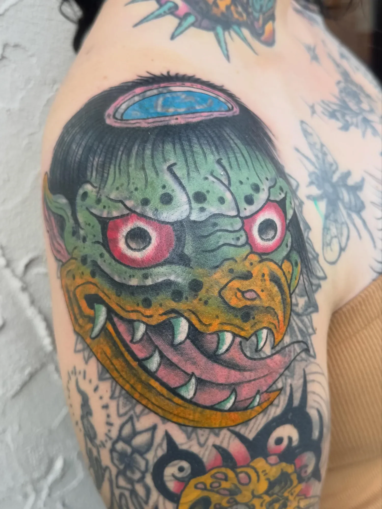  Kappa — green water demon with red eyes, yellow beak, and plate on head. Custom yokai piece on shoulder cap, built to flow with the deltoid. Two angles showing wrap.
