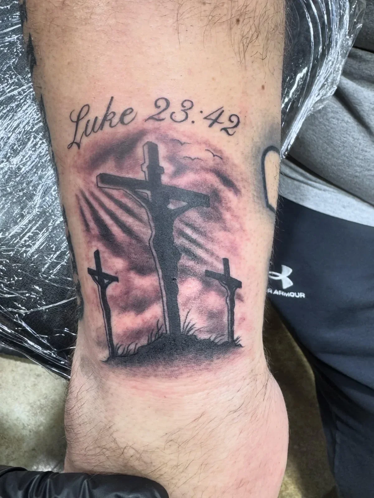 Powerful religious tattoo depicting three crosses at Calvary silhouette against cloudy sky with rays of light, script reading Luke 23:42. Black and gray Christian tattoo by Brett Semrick.