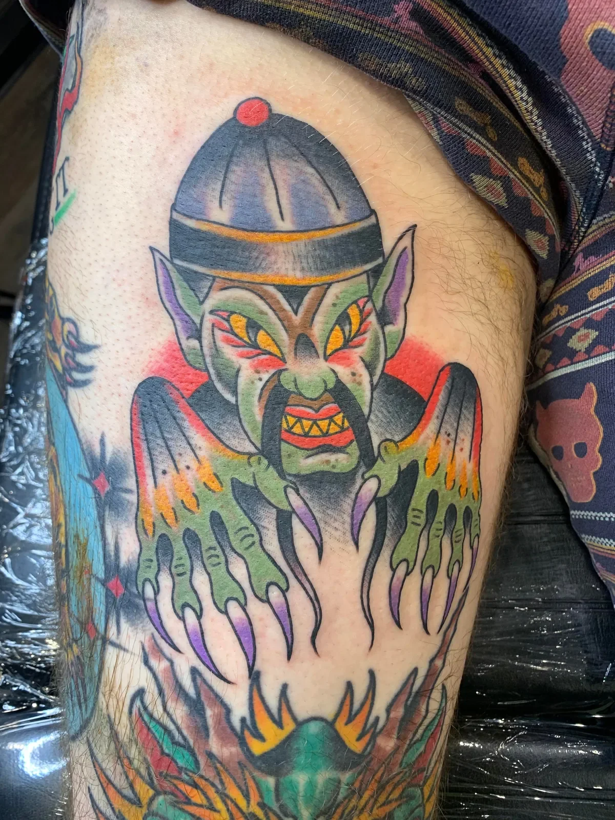 Colorful tattoo of a demonic creature with green skin, purple ears, fierce eyes, and a sinister expression, wearing a helmet and with bat-like wings extending from its back.