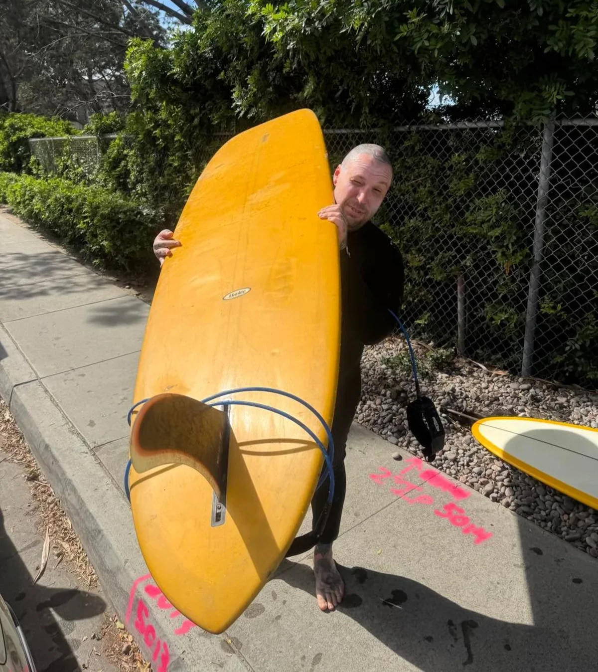 A man holding a yellow surfboard on a sidewalk with a chain-link fence and green bushes in the background.