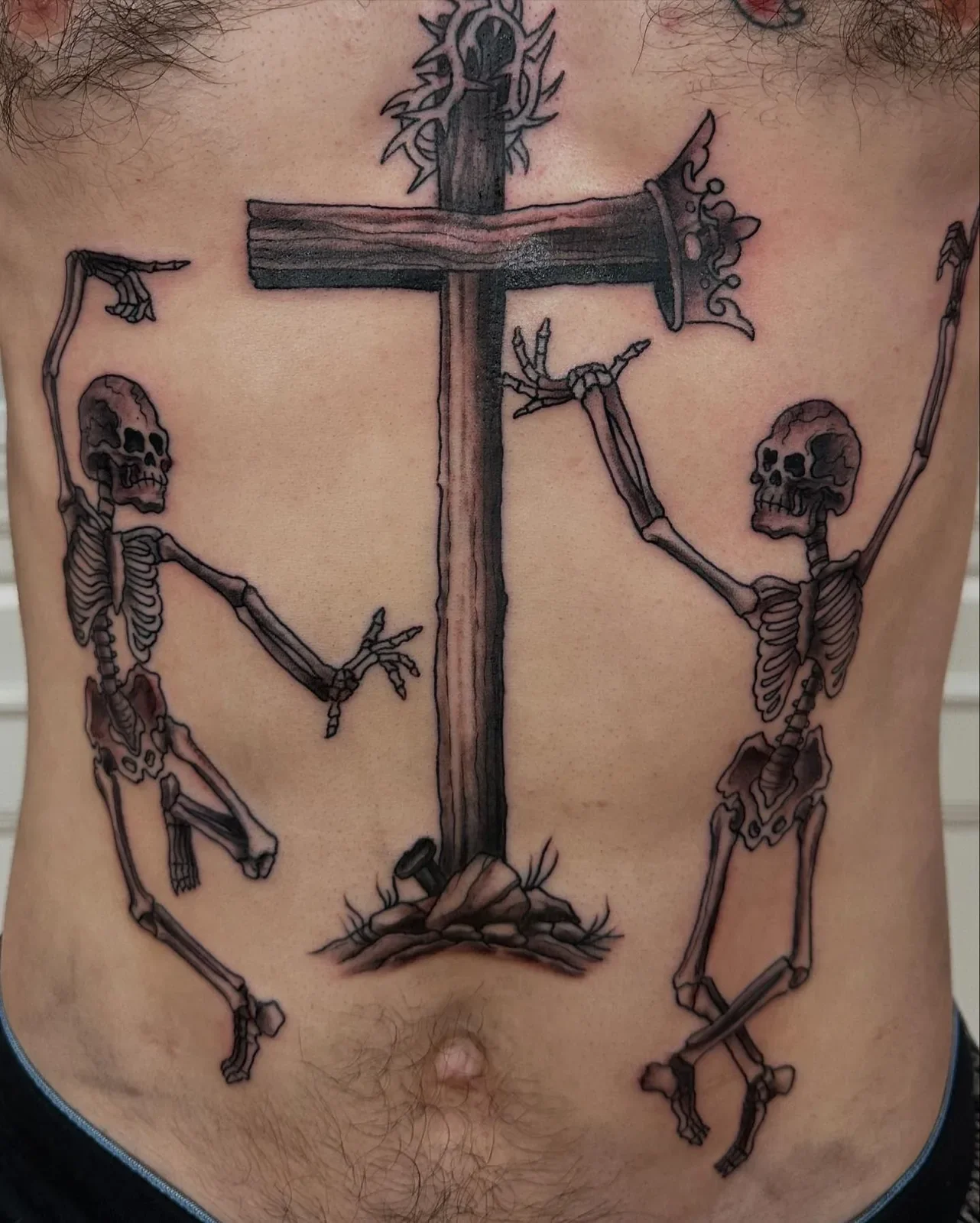 Unique dark religious tattoo with wooden cross wearing crown of thorns and crown, flanked by two dancing skeletons reaching up. Black and gray illustrative piece by Brett Semri