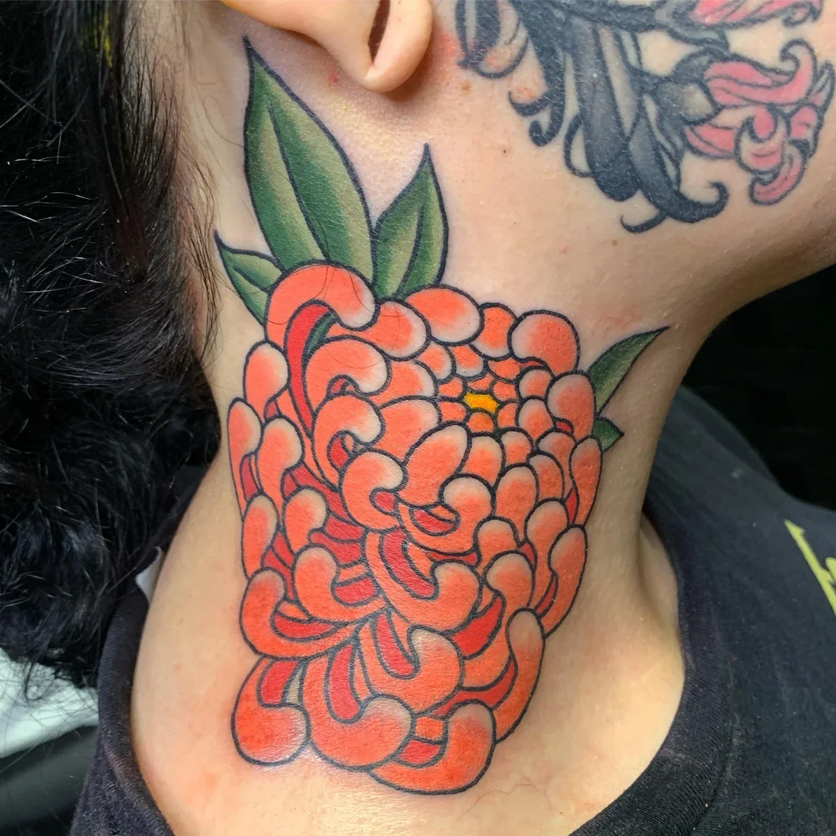 Tattoo of an orange chrysanthemum flower with green leaves on a person's neck.