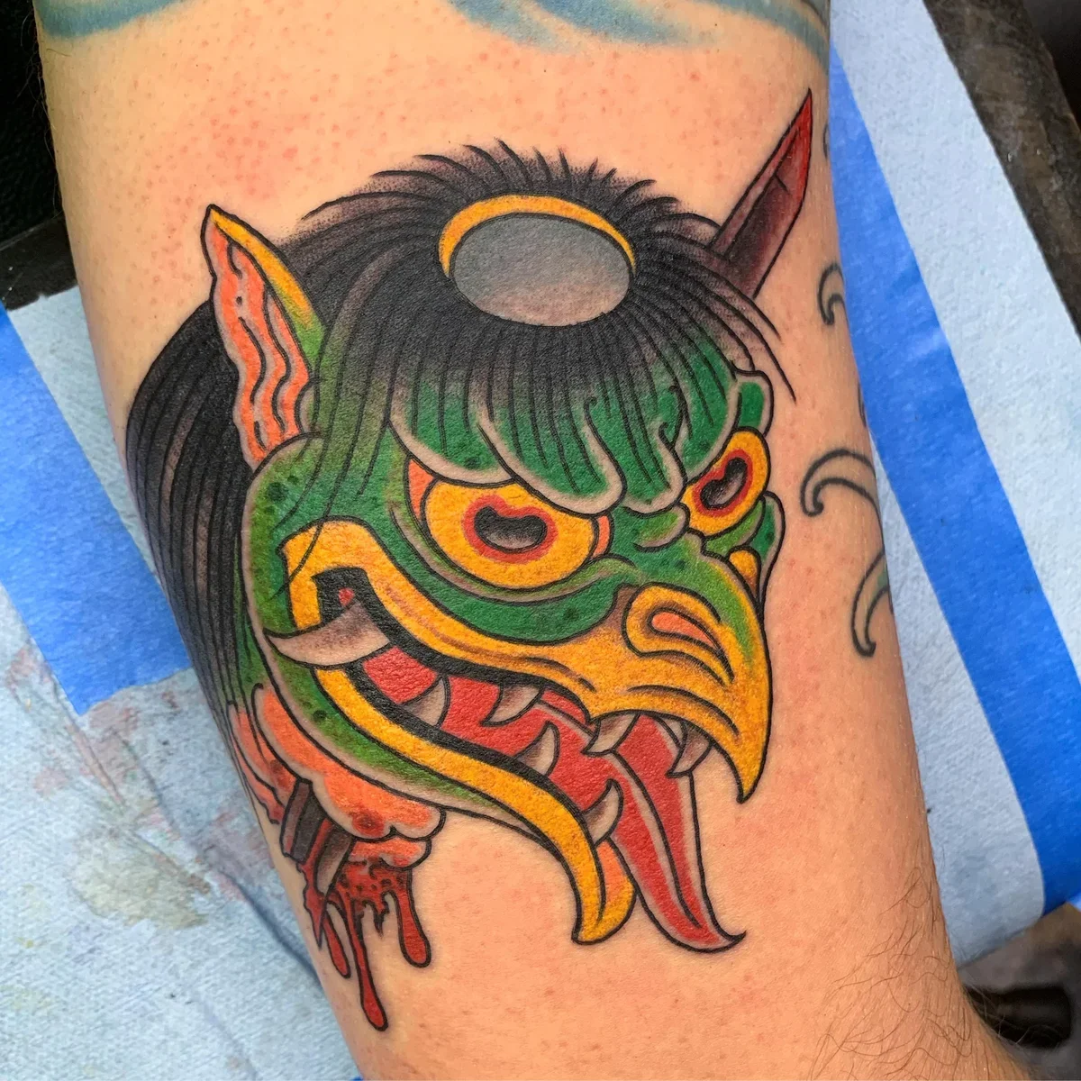 Close-up of a colorful tattoo featuring a demon or ogre face with sharp teeth, red tongue, green skin, and a horn on the forehead, with a blue background.