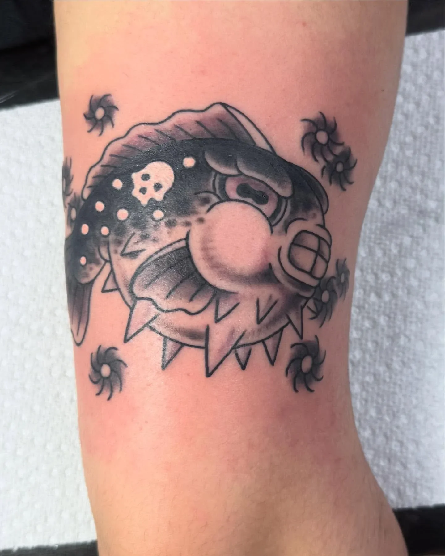 Black and gray tattoo of a cartoonish angry fish with sharp teeth, surrounded by small swirling designs on skin.