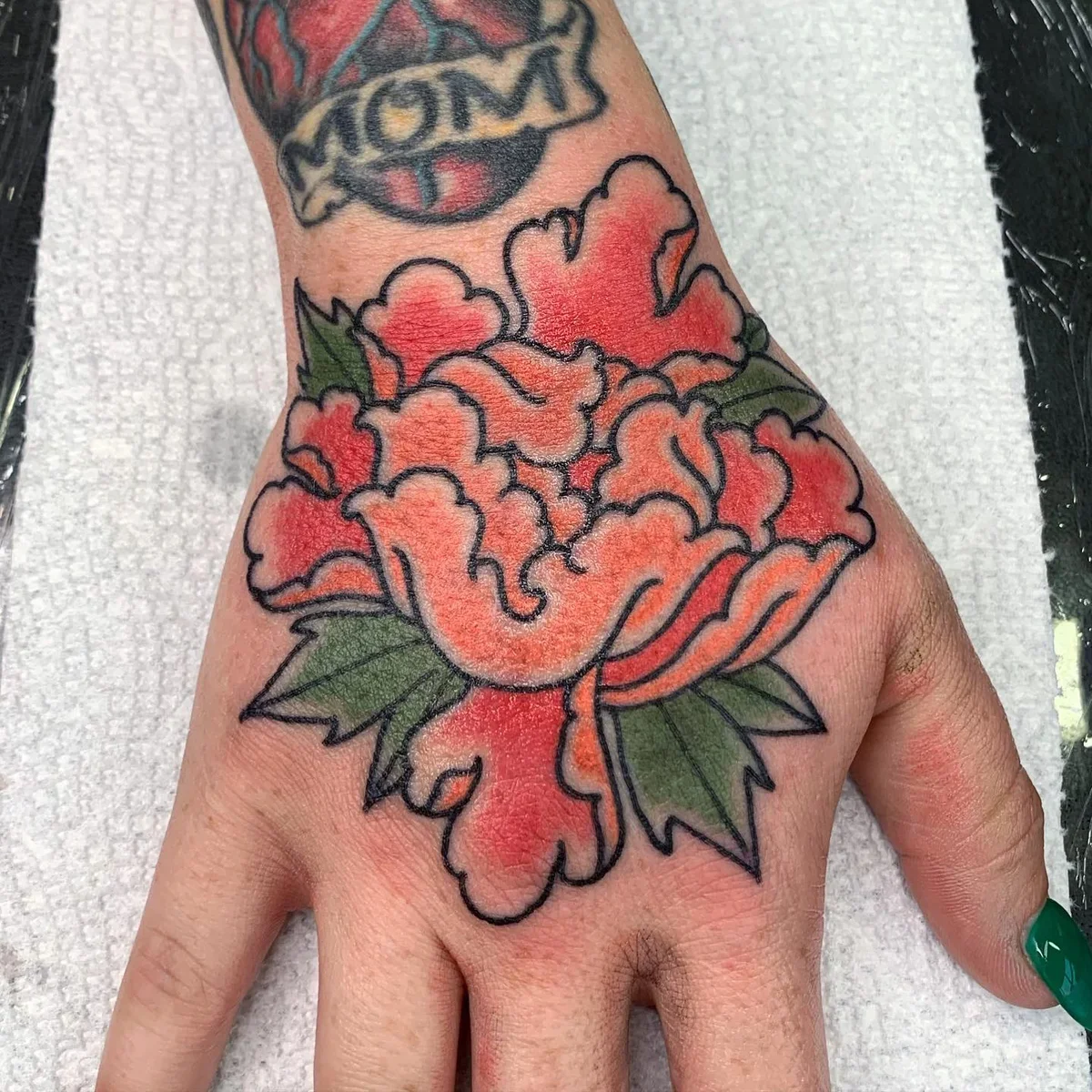 Colorful tattoo of a pink and orange peony flower with green leaves on a person's hand.