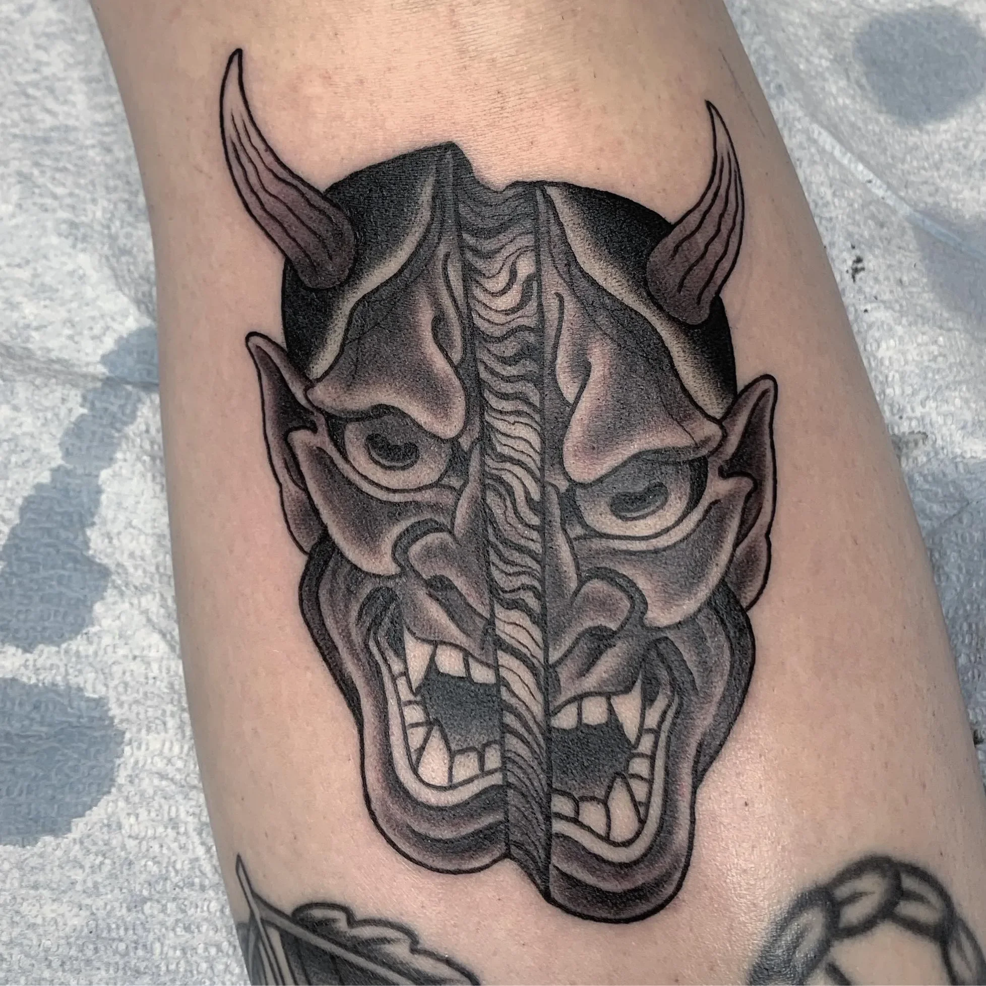 Split Hannya — half dark, half light with a wood grain / leaf pattern center seam. Black and grey with whip shading and smooth gradations. Lower leg piece, bold and graphic.

