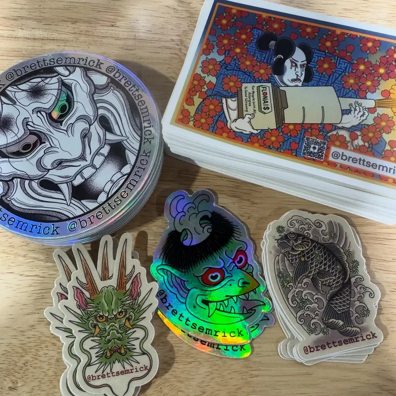 Collection of stickers and trading cards on a wooden surface, including a holographic sticker of a colorful demon, a black and white tiger sticker, a dragon sticker, and a stack of illustrated tarot cards. The circular sticker features a detailed bla