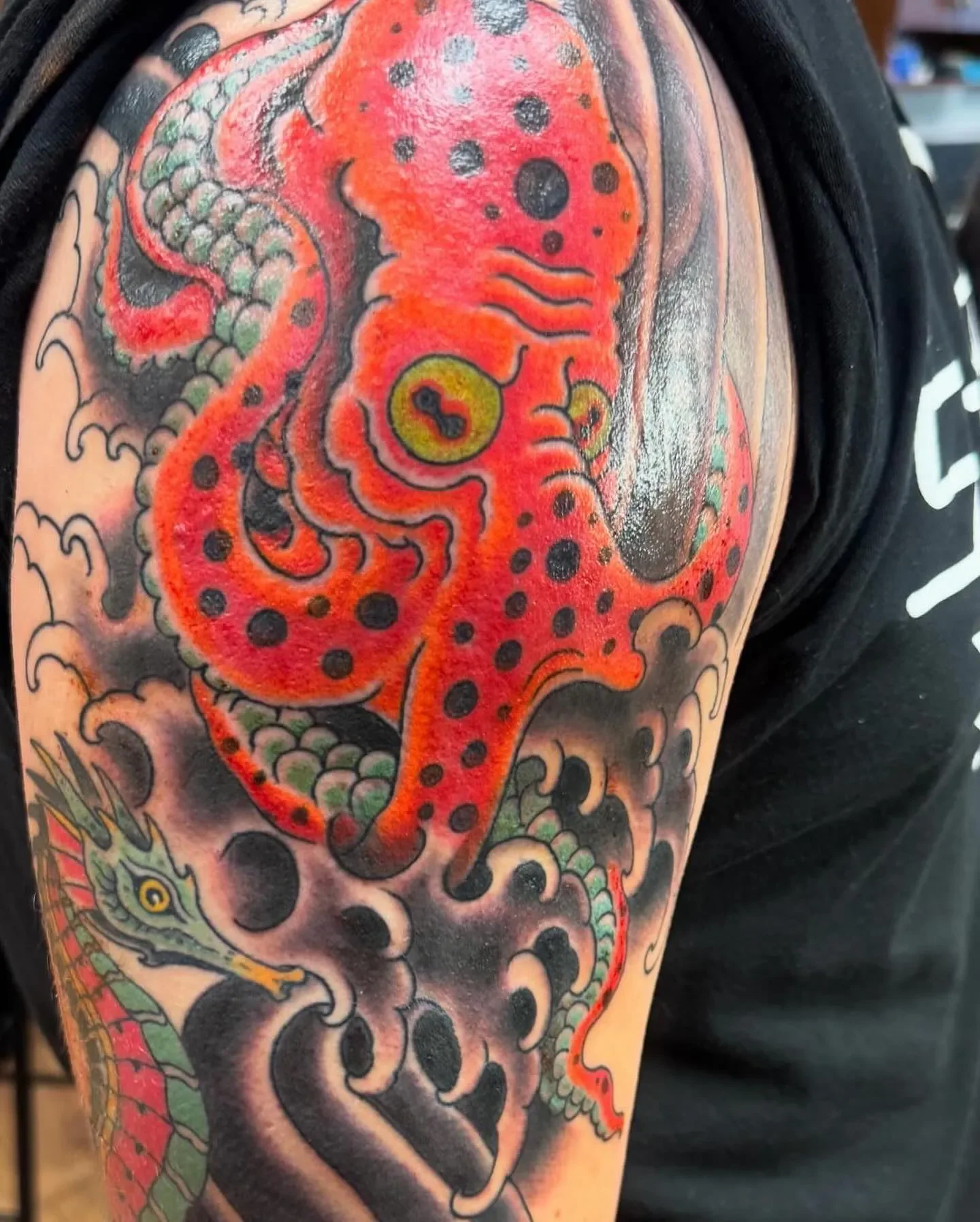  Tako — bright red octopus with black spots, green under-scales and yellow eyes, flowing into a green seahorse below. Bold traditional color with heavy black smoke fill. Shoulder piece.
