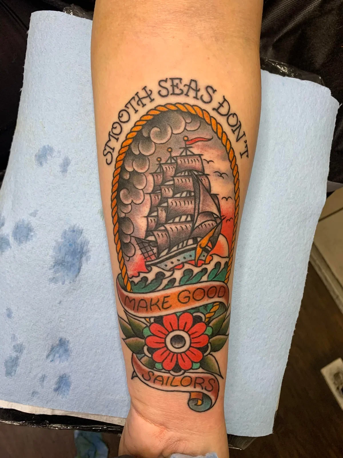 Clipper ship in stormy seas framed by rope border, scroll banners and red flower — classic motivational sailor tattoo.

