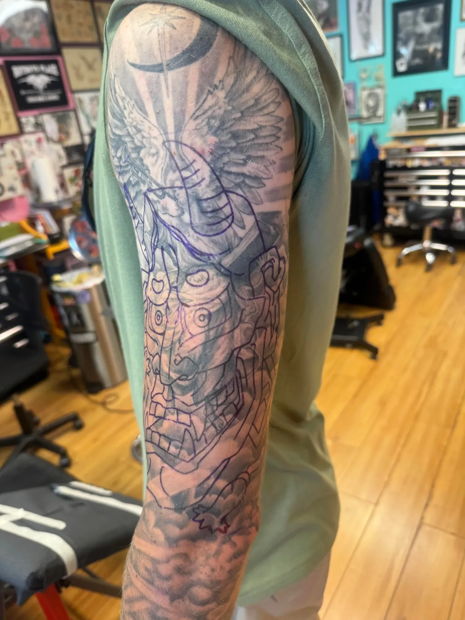 A person's arm with a detailed tattoo of an eagle with wings spread, drawn partly in black ink with the design outlines visible, in a tattoo shop.