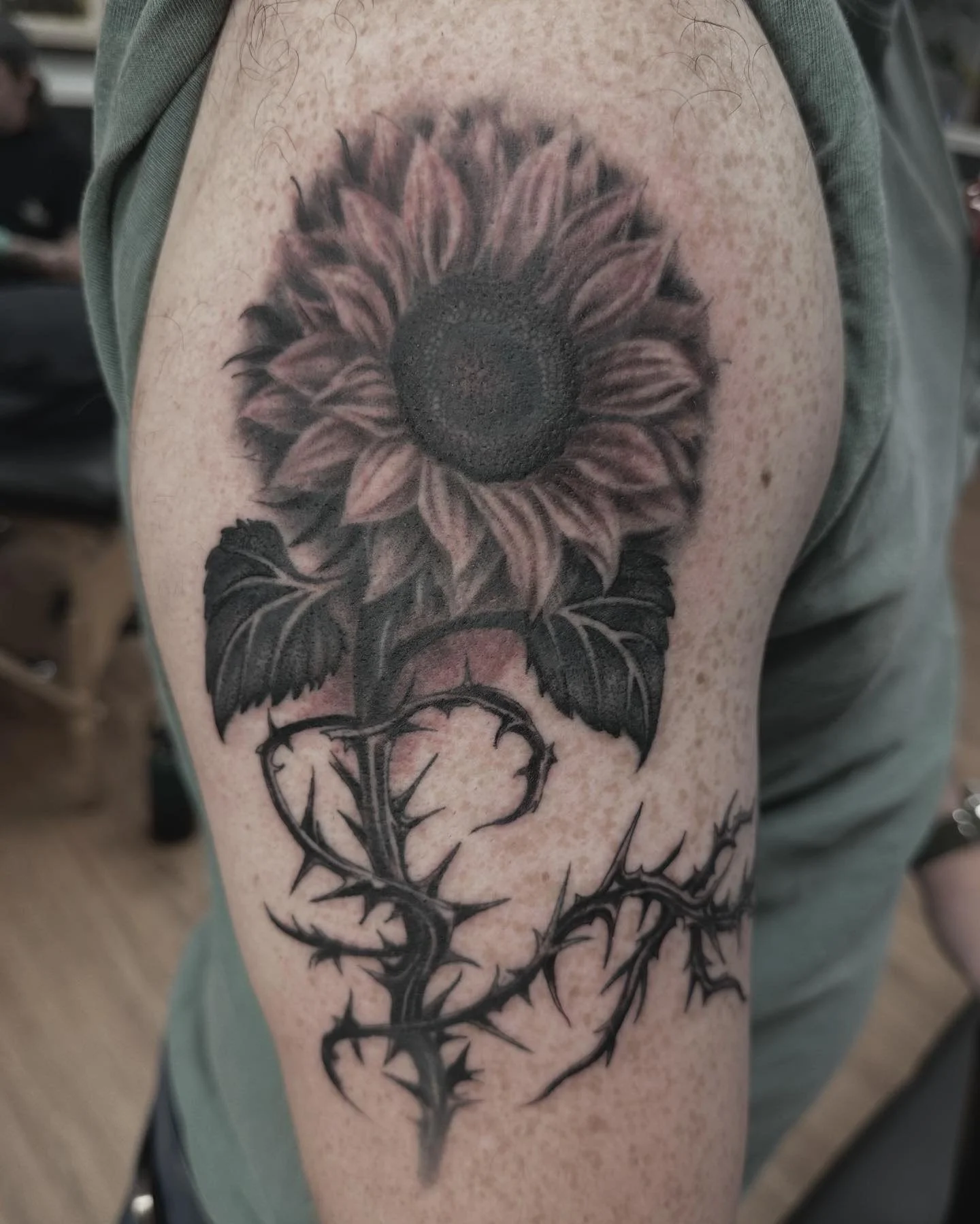 Sunflower with dark leaves and thorn-vine stem wrapping around arm. Illustrative blackwork with realism flower head.
