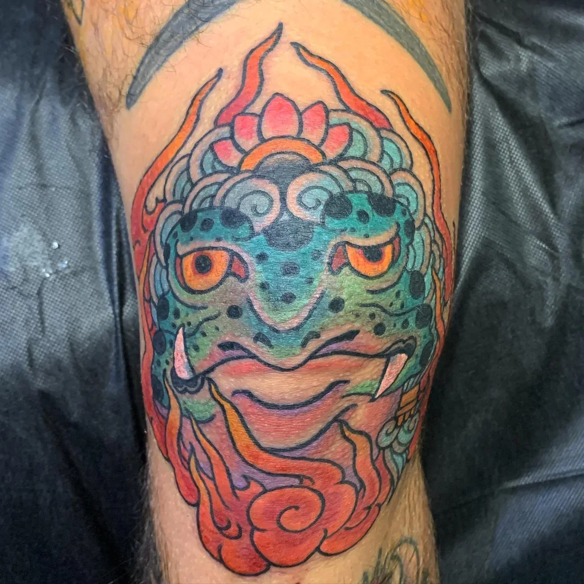 Colorful tattoo of a lizard or gecko with vibrant patterns on the face, surrounded by flames and decorative elements.