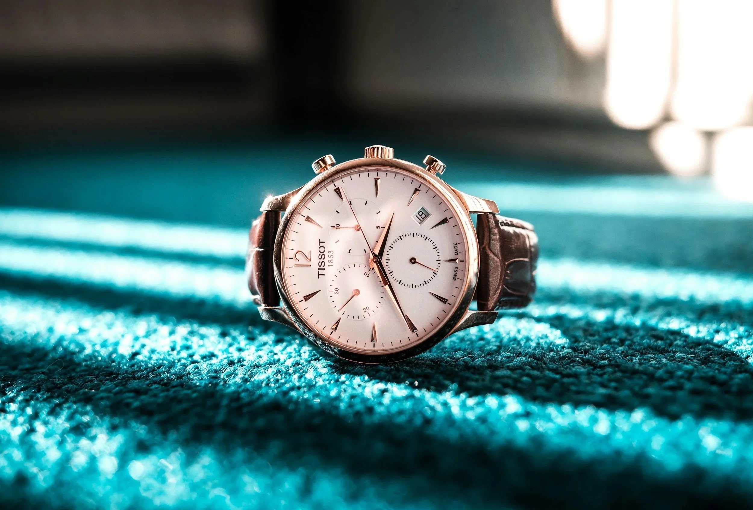 Tissot tradition chronograph dress watch with brown leather straps laid on a teal or turquoise rug carpet