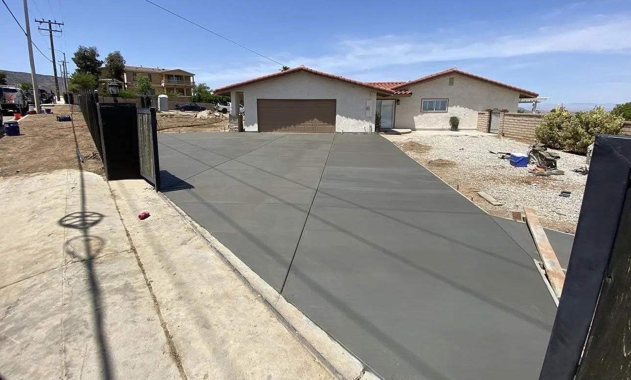 Newly poured concrete driveway leading to a house with a two-car garage, side yard with gravel and plants, and surrounding fencing on a sunny day.