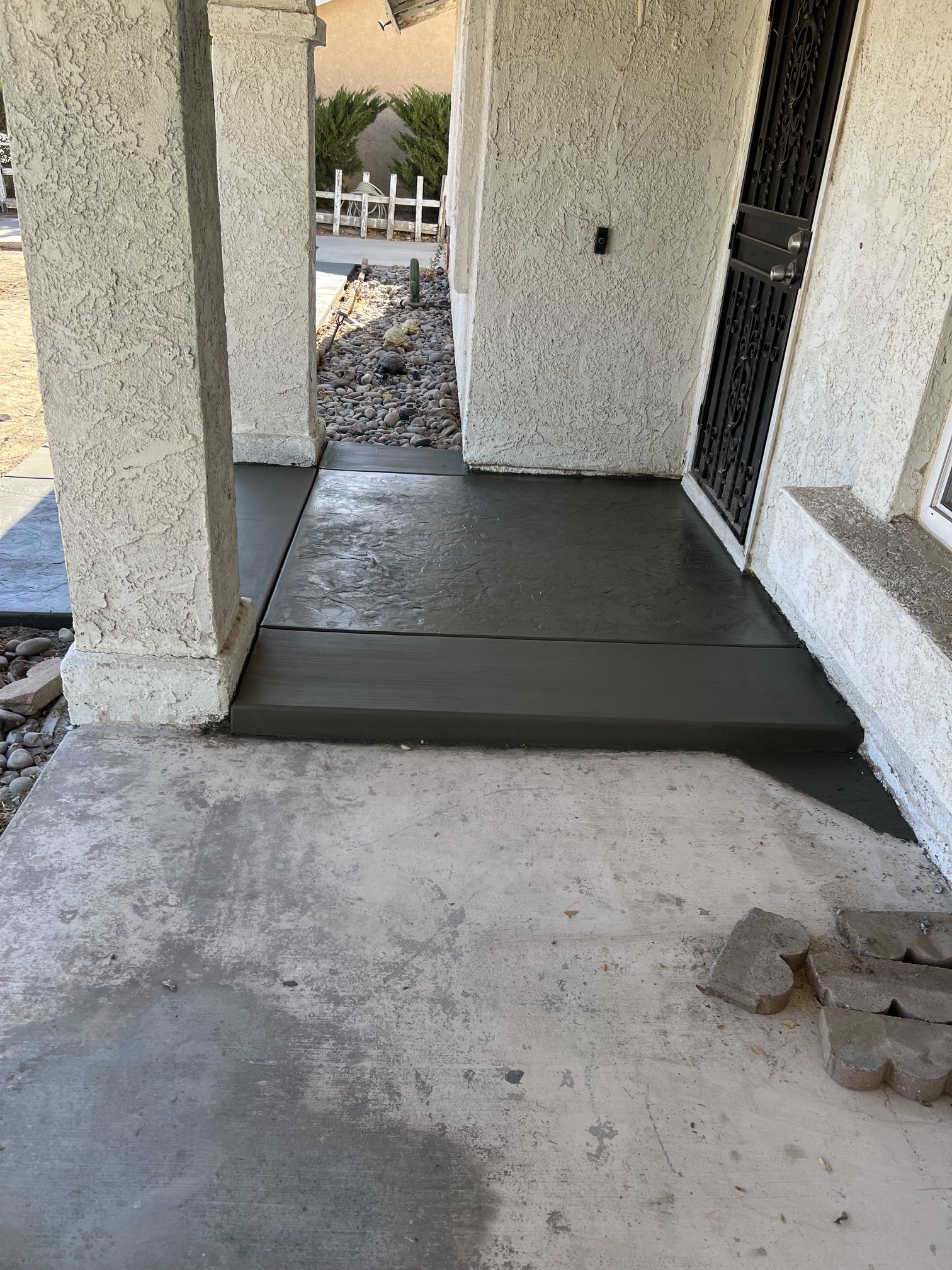 Front porch with concrete floor, textured white stucco walls, a metal security door, and a small garden area with rocks and plants visible through an opening.