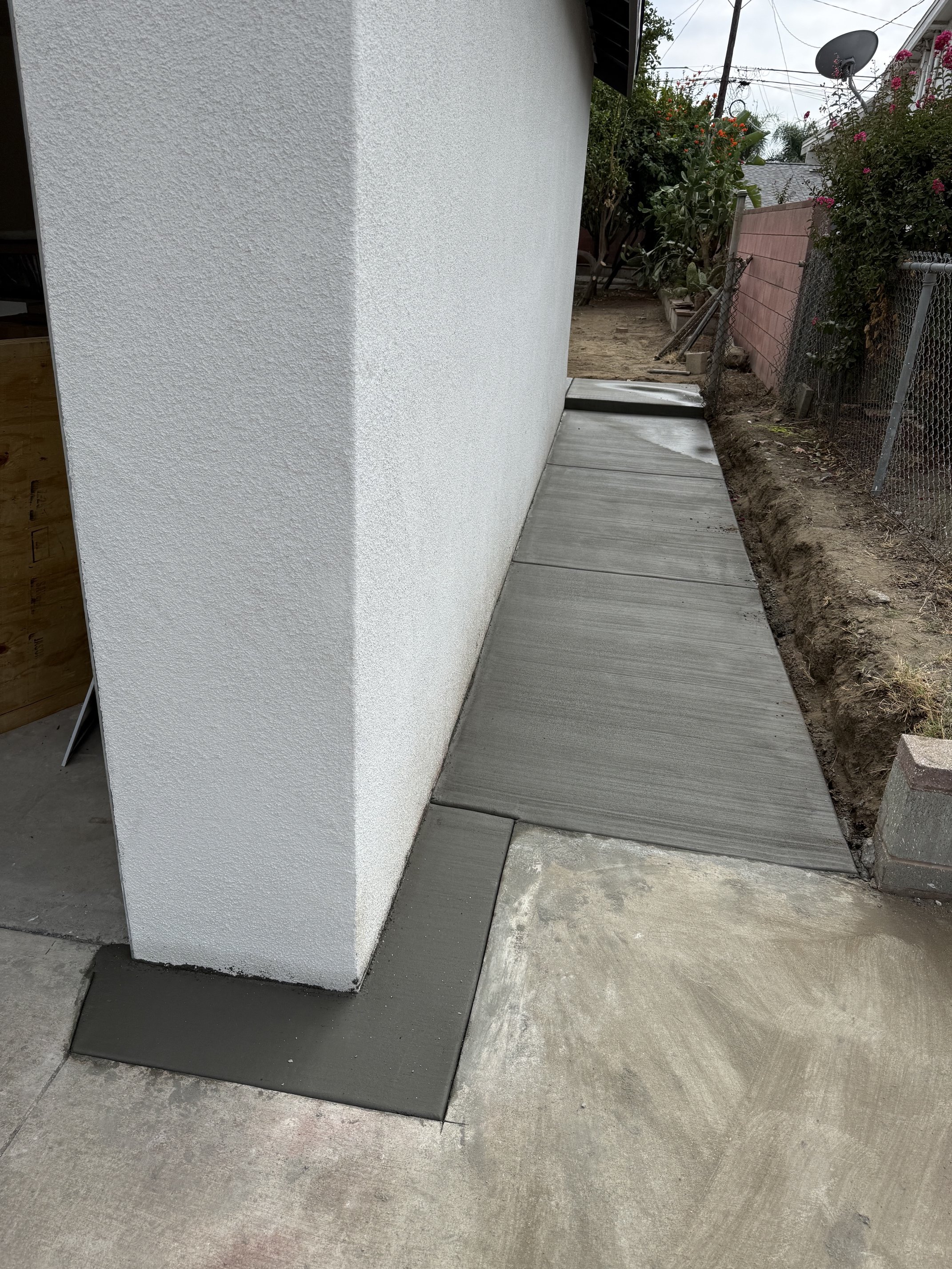 Newly poured concrete sidewalk alongside a white textured exterior wall of a building, with an adjacent dirt area and a neighboring fence with plants, logs, and a satellite dish in the background.