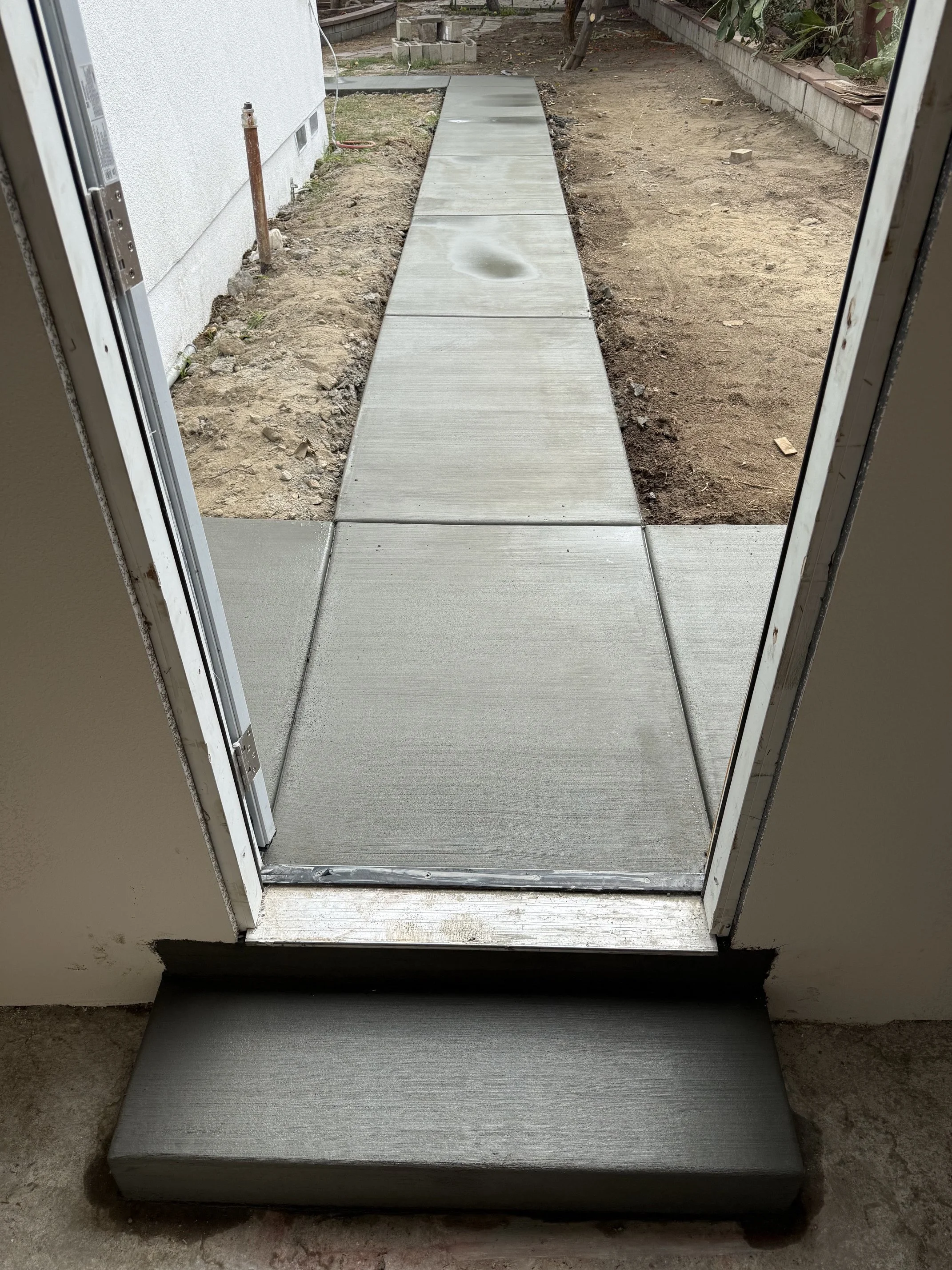 Newly installed concrete walkway outside a house, viewed from inside. The walkway extends from the door into a yard with dirt and some landscaping.
