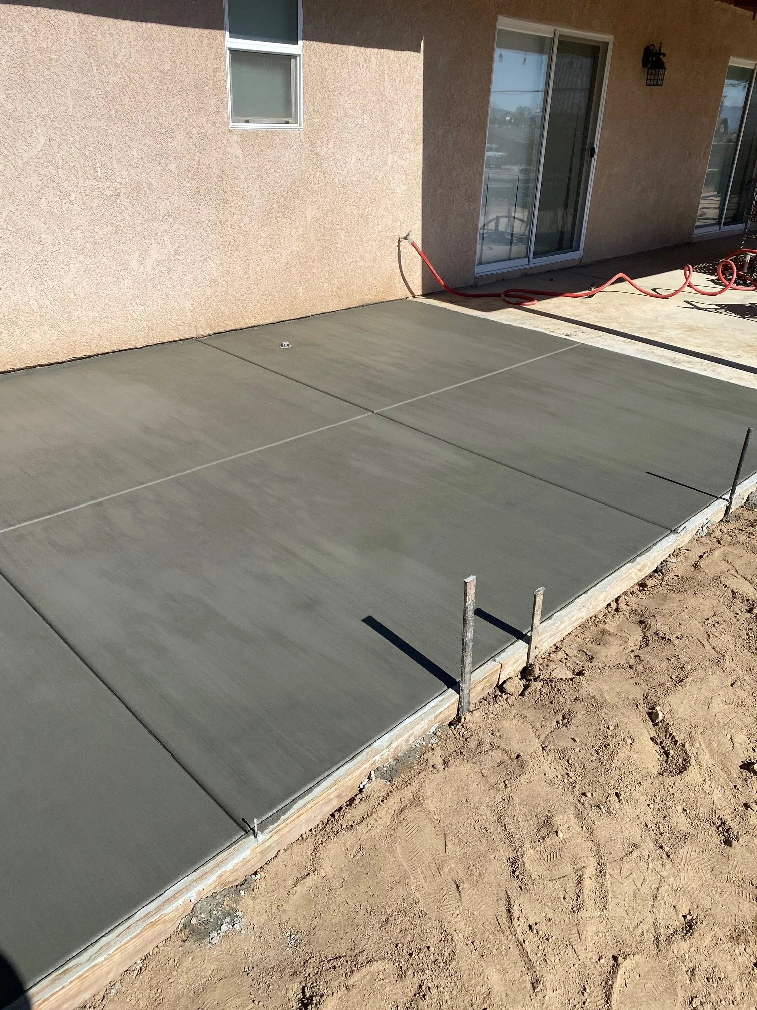 Concrete patio being poured outside a house with two sliding glass doors and a window, with construction tools and materials visible.