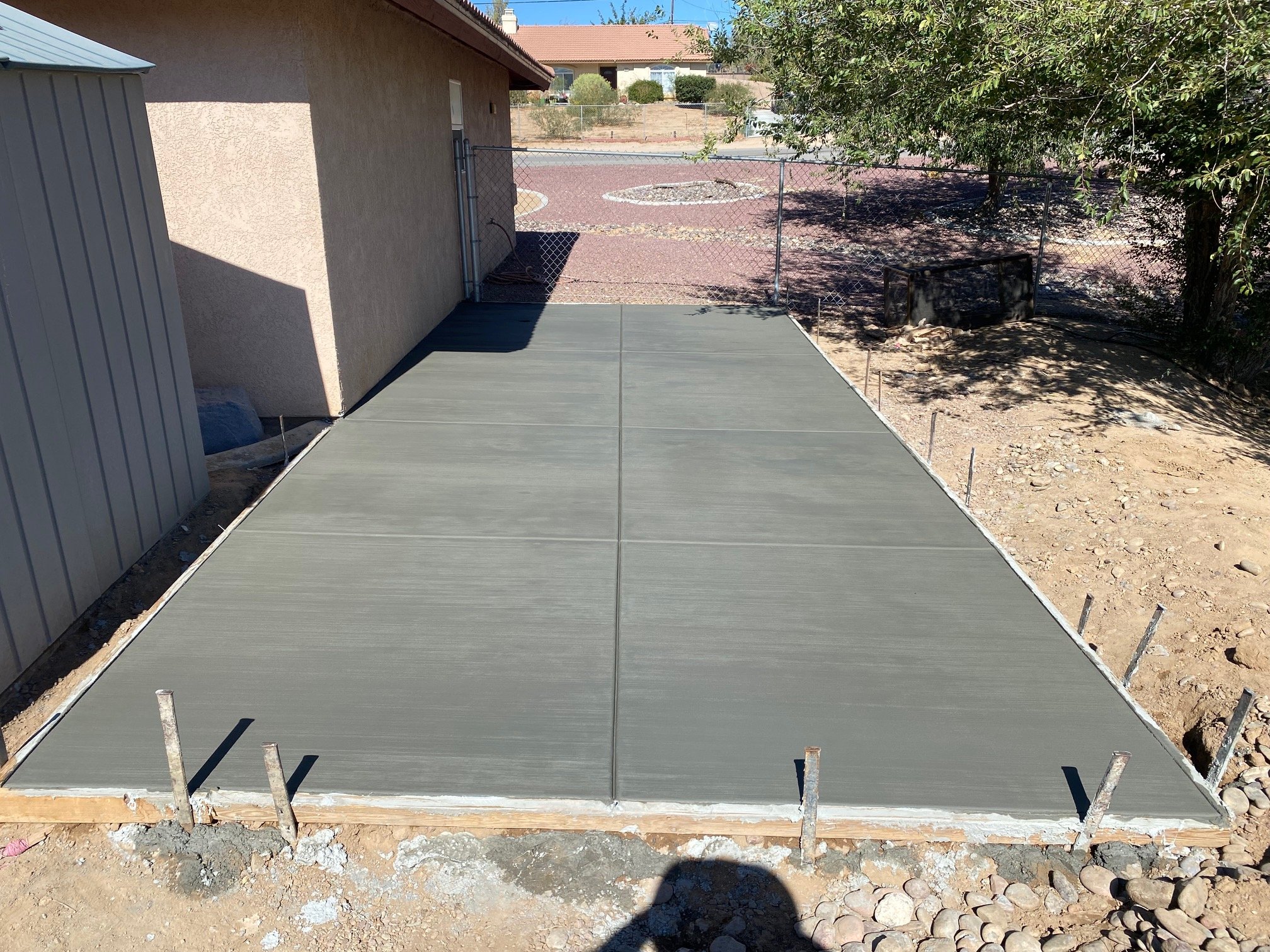 A concrete patio area under construction with a partially completed strip of concrete. There is a chain-link fence in the background and trees and houses beyond it. Shadows of trees and a person are cast onto the ground.