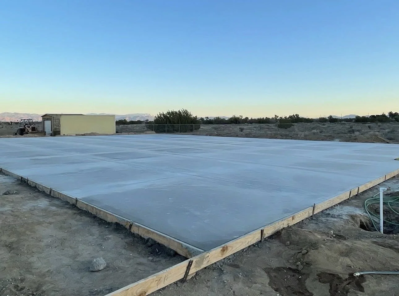A flat concrete slab on a construction site with a dirt ground, a small building in the background, and a clear sky at dusk.