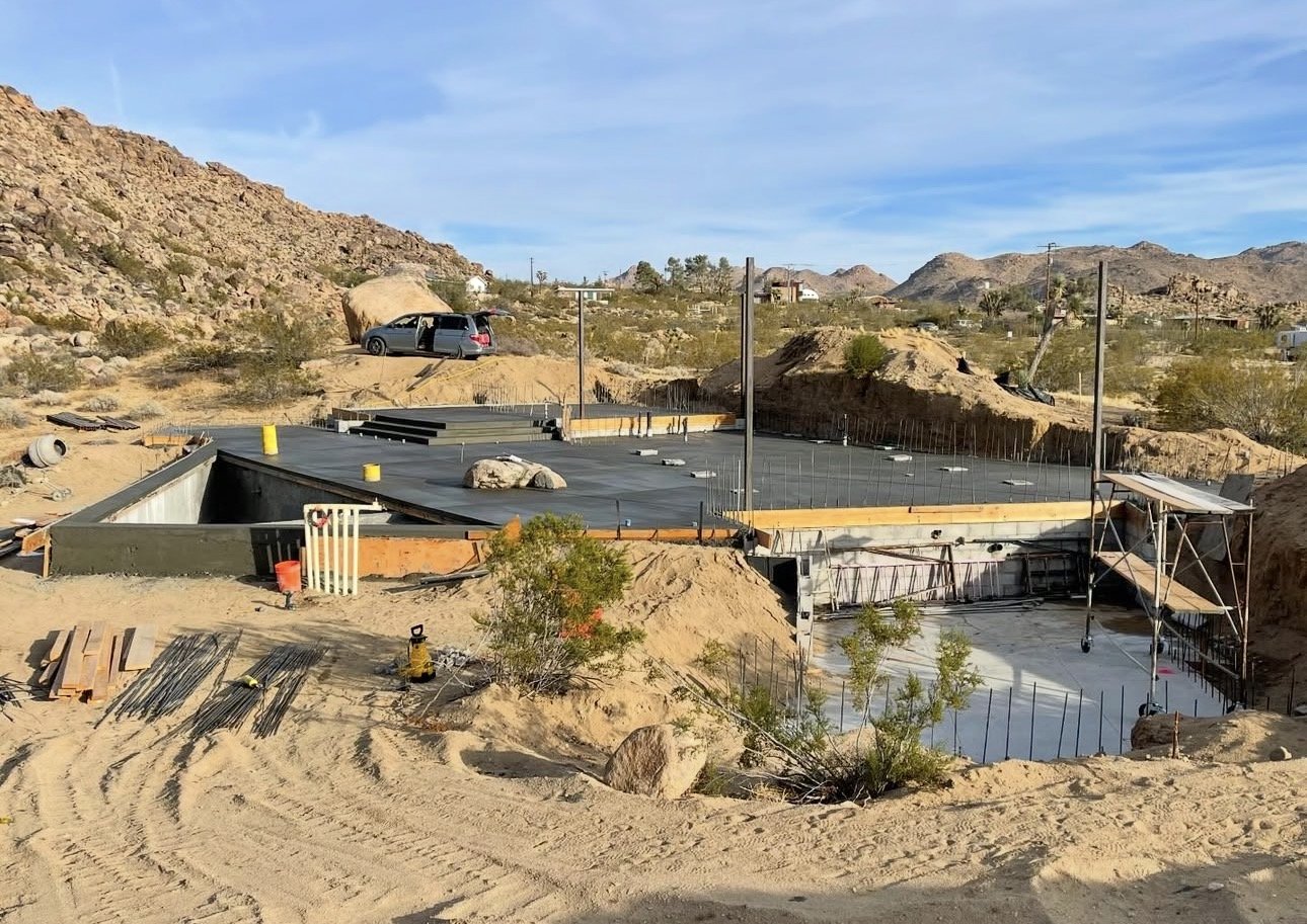 Construction site in a desert area with a building foundation being prepared, surrounded by sand and rocks, with mountains and a blue sky in the background.