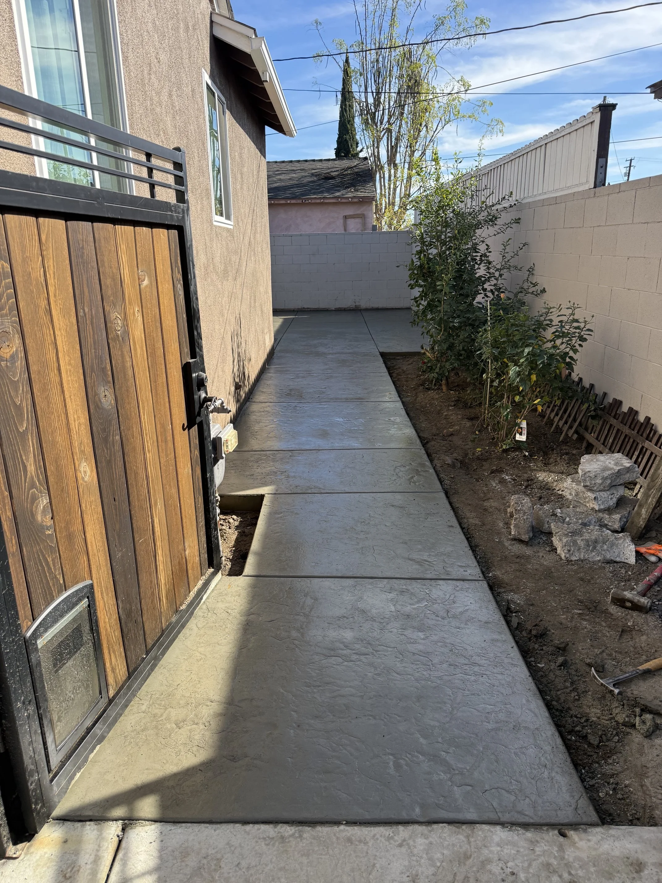 A freshly poured concrete sidewalk next to a house with a wooden gate on the left. A white brick wall and greenery are on the right, with some rocks and tools in the dirt. The sky is clear and blue.