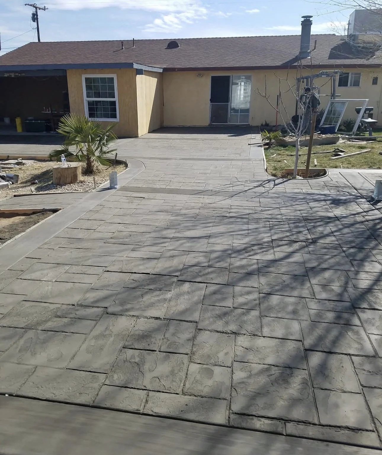 A backyard patio under construction with newly installed concrete pavers, adjacent to a house with unfinished siding and small trees, some construction materials, and tools visible.