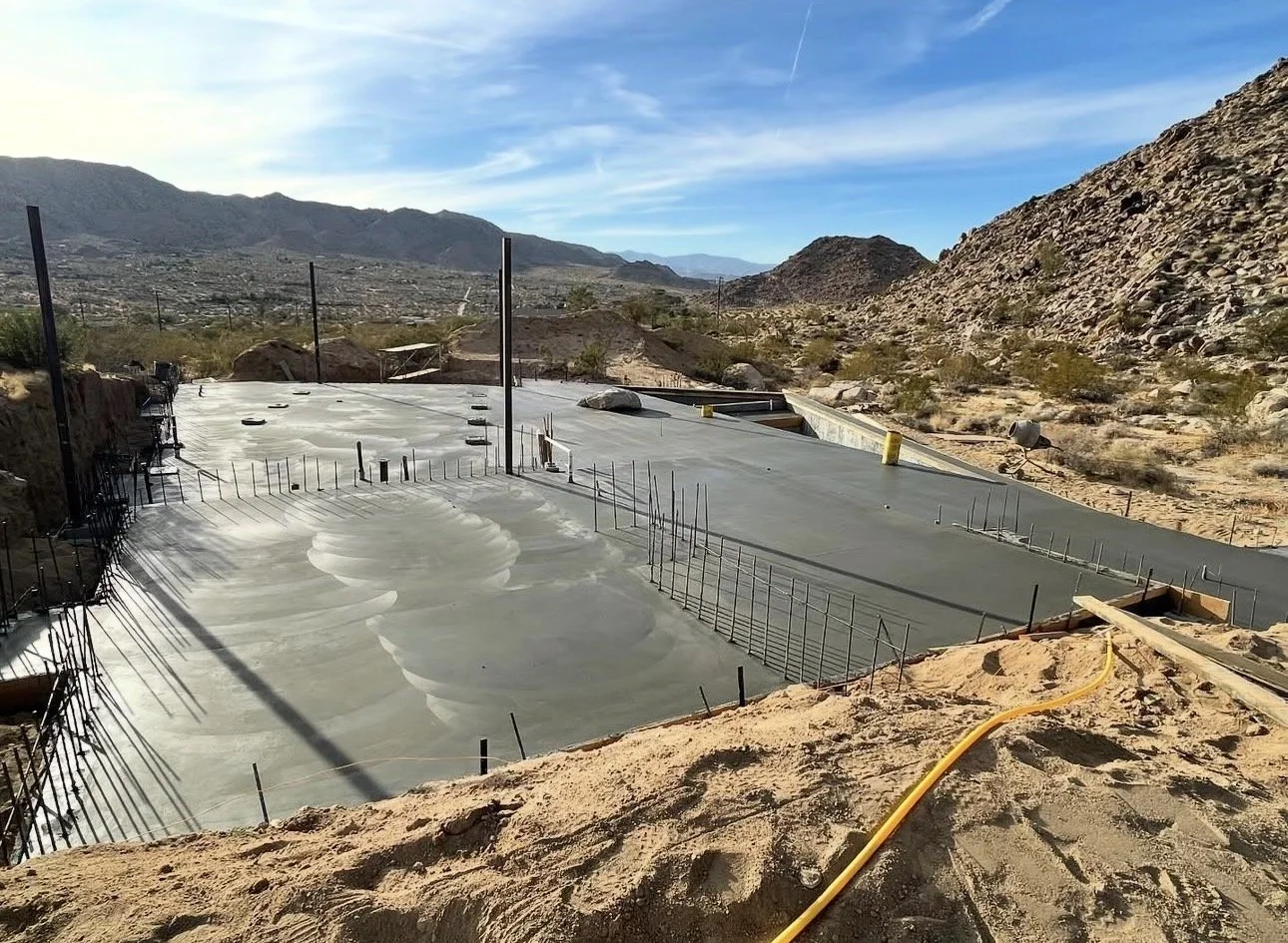 Construction site of a swimming pool in a desert landscape with mountains in the background, concrete shell being formed, and construction materials and tools around.
