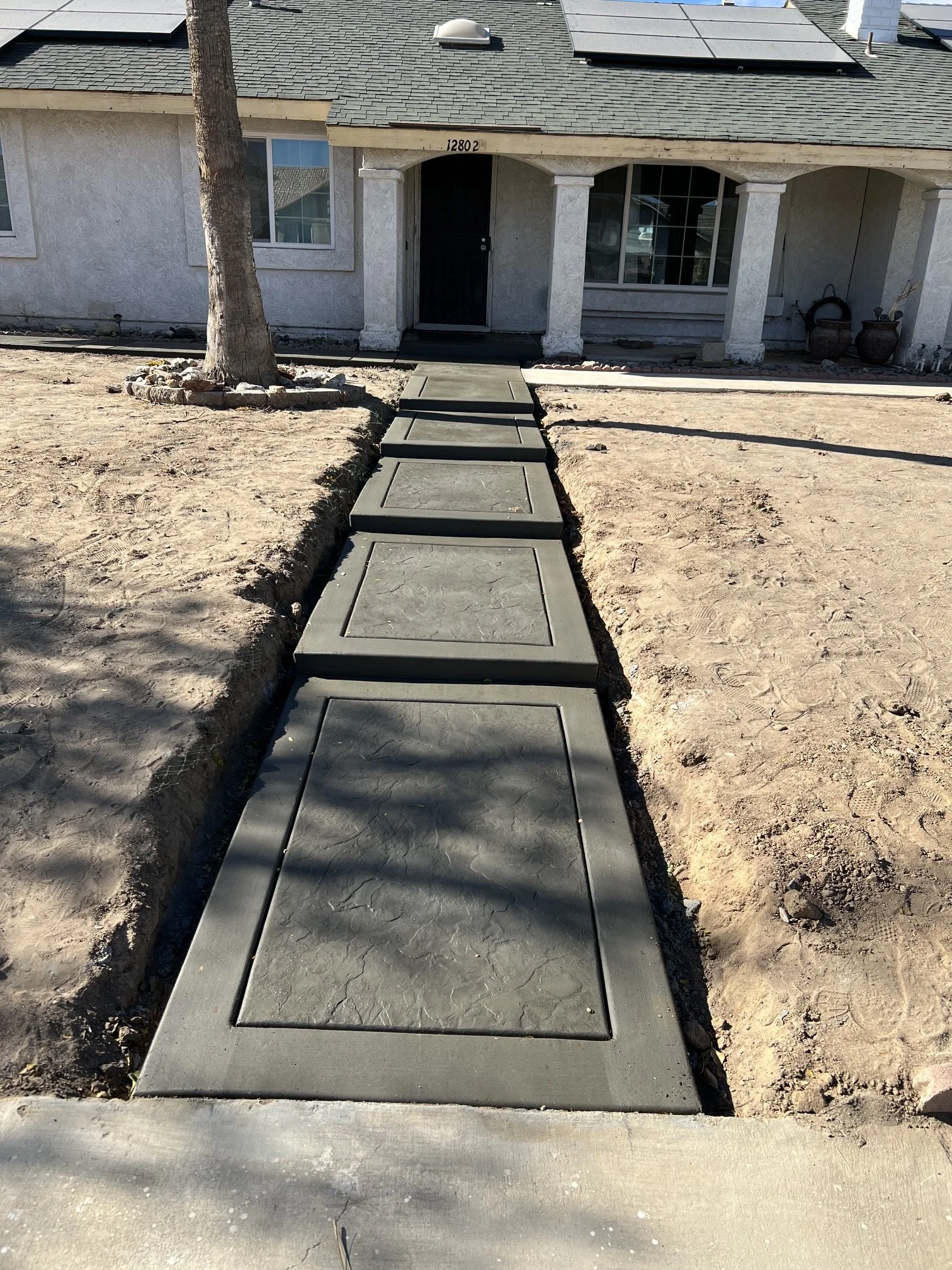 Newly installed concrete sidewalk leading to the front door of a house. The house has a tiled roof, large windows, and a covered porch. The area surrounding the sidewalk is bare dirt.