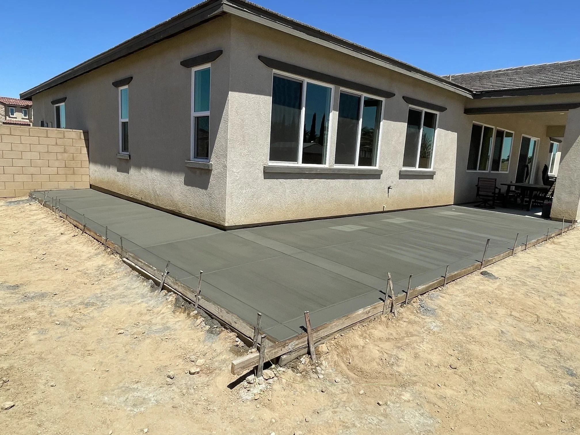 Newly poured concrete patio with wooden framing around the edges, adjacent to a beige house with multiple windows.