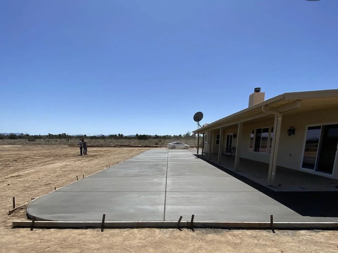 Newly poured concrete driveway next to a house with a covered patio, satellite dish, and large windows, set in a rural area with a clear blue sky.