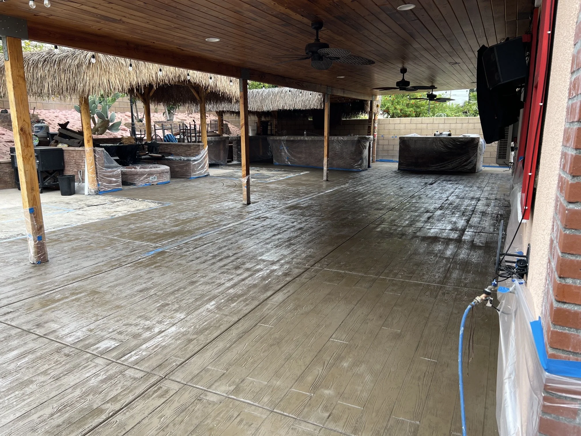 Outdoor covered patio area with wooden ceiling, ceiling fans, and brick wall. The floor appears to be freshly stained or sealed, with visible protective tape around some areas. There are tiki-style straw umbrellas, and some patio furniture covered wi