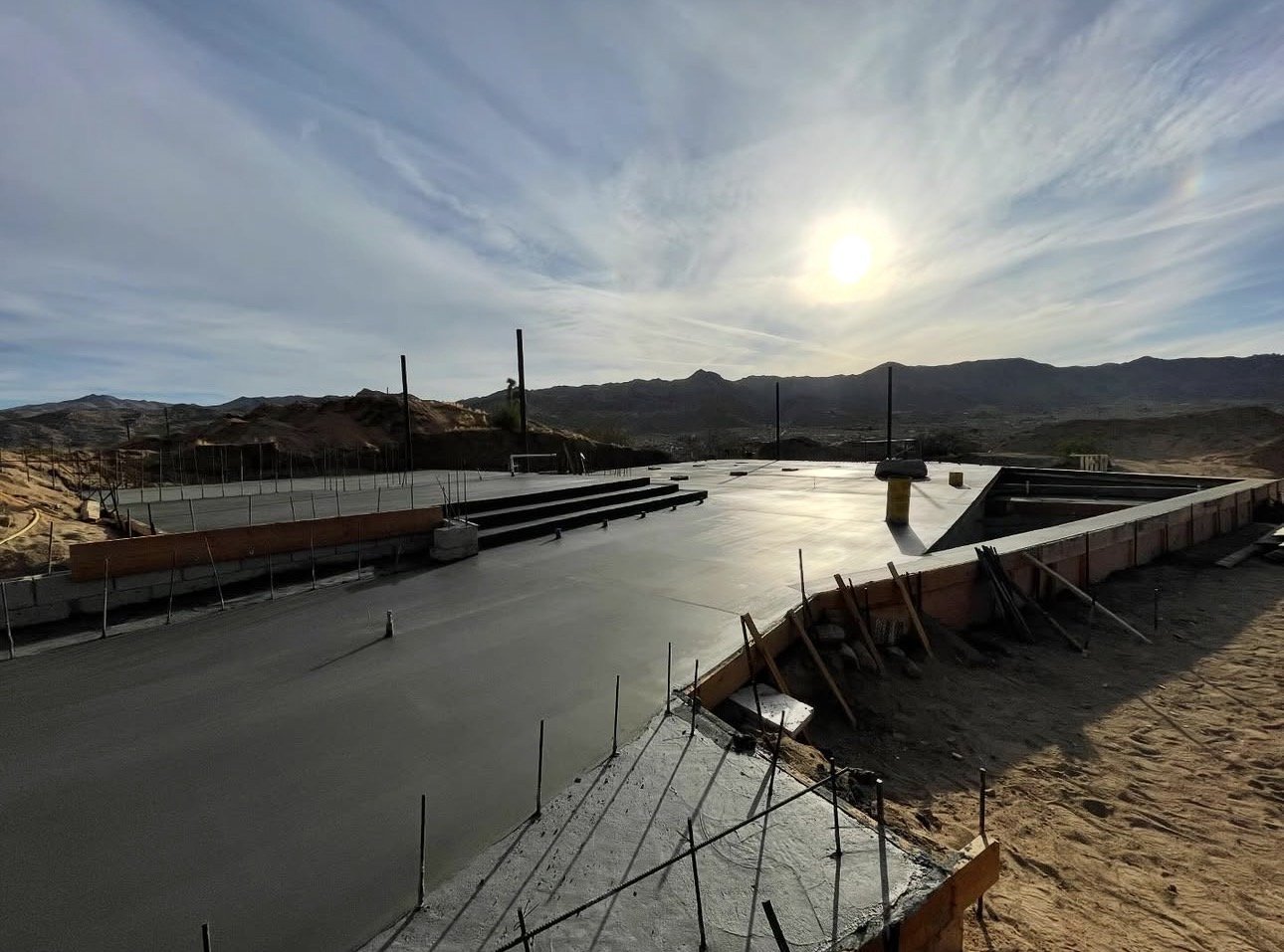 Construction site of a concrete foundation with rebar and wooden formwork, set against a desert landscape with distant mountains and a partly cloudy sky.