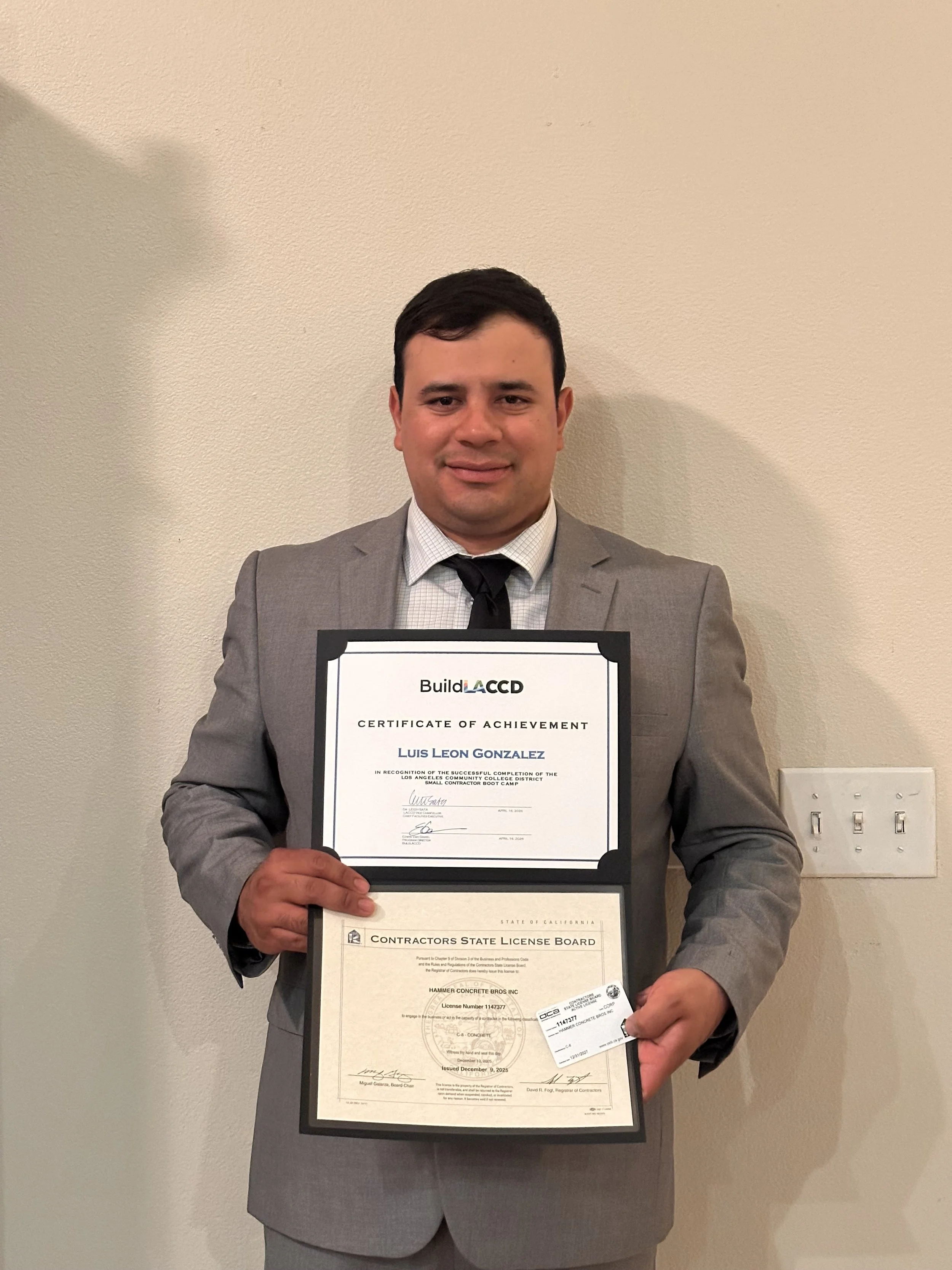 Hammer Concrete Bros Inc CEO holding a certificate and a slash card in front of a plain beige wall.